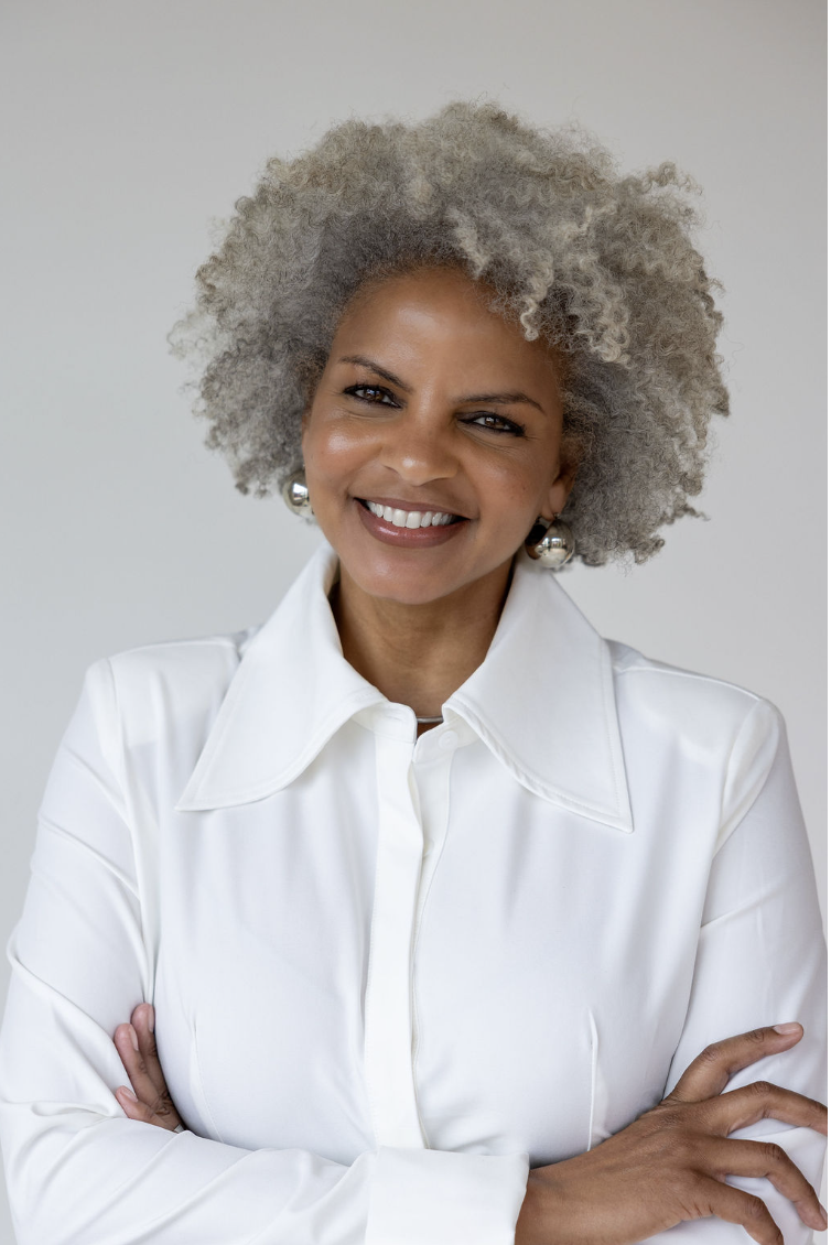 Smiling middle-aged woman with gray curly hair, wearing a white blouse and silver earrings, standing with arms crossed against a plain background.