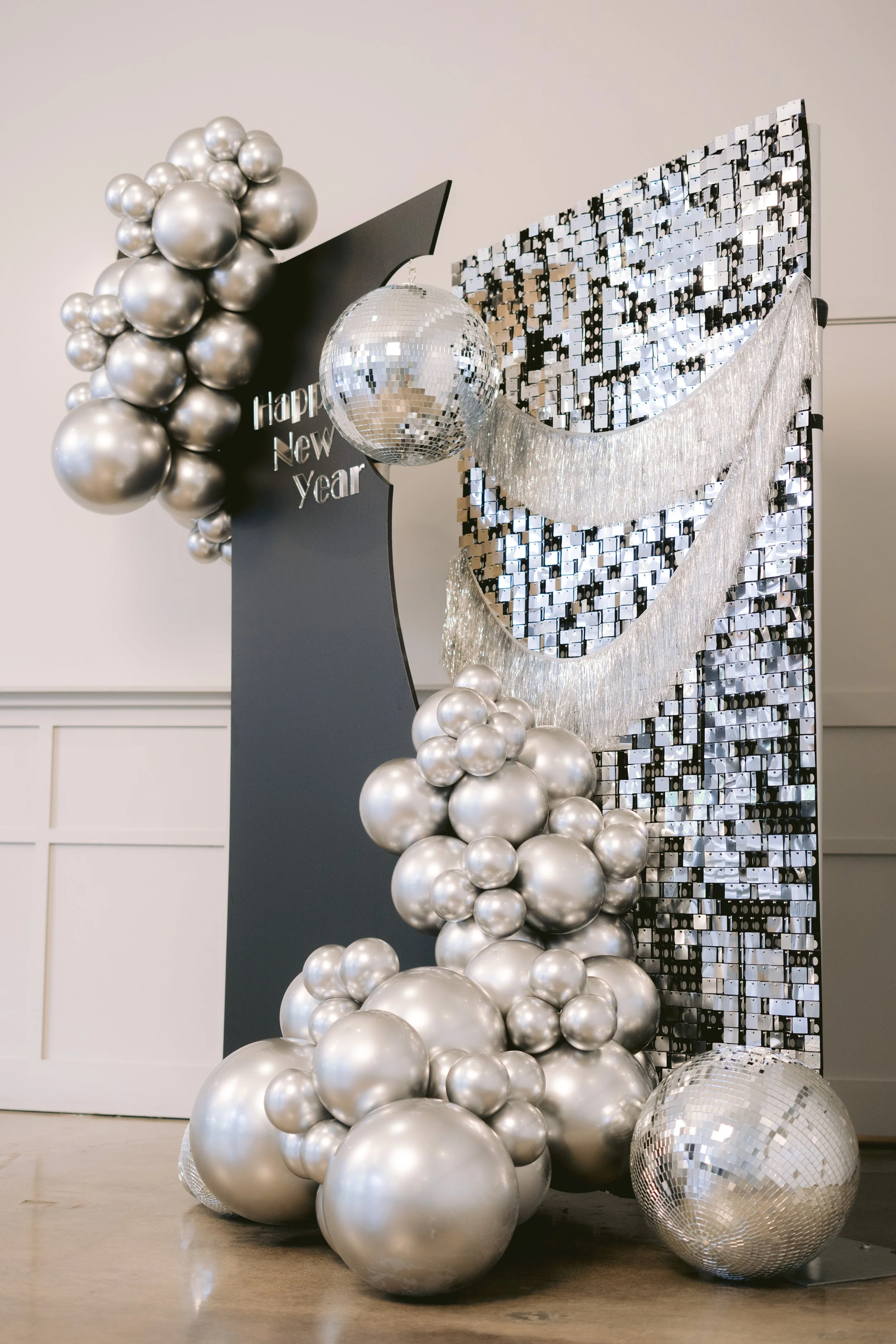 Decorative New Year celebration setup with silver balloons, a disco ball, and a black sign reading 'Happy New Year' in front of a mirrored backdrop.