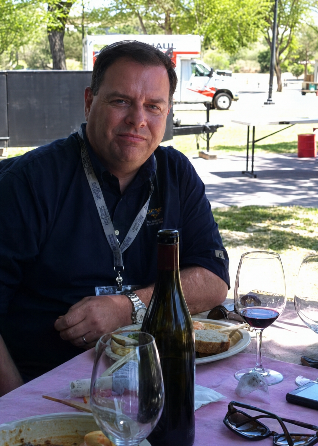 Two men at a table with champagne bottles and empty wine glasses, one man is sitting and the other is standing and opening a bottle, with a plain neutral wall in the background.