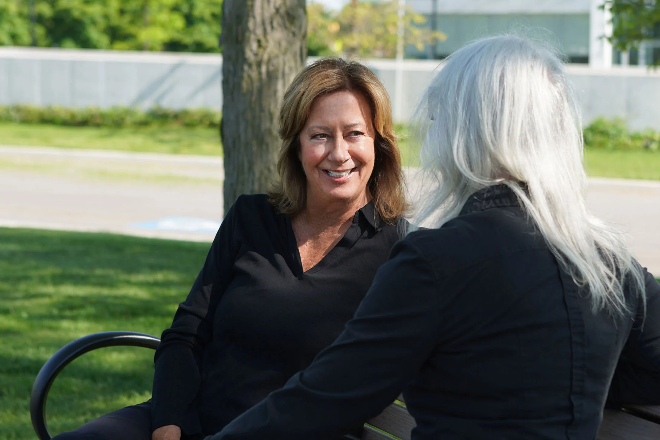 Two women engaged in a conversation outdoors on a bench in a park with green trees and grass in the background.