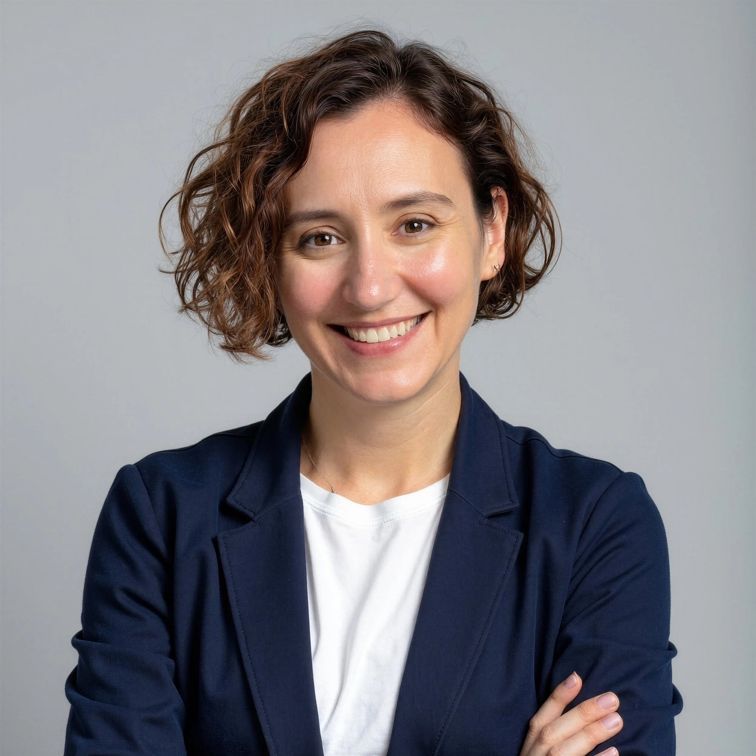 Close-up of a smiling woman with short, wavy brown hair wearing a navy blazer and white t-shirt, against a plain gray background.