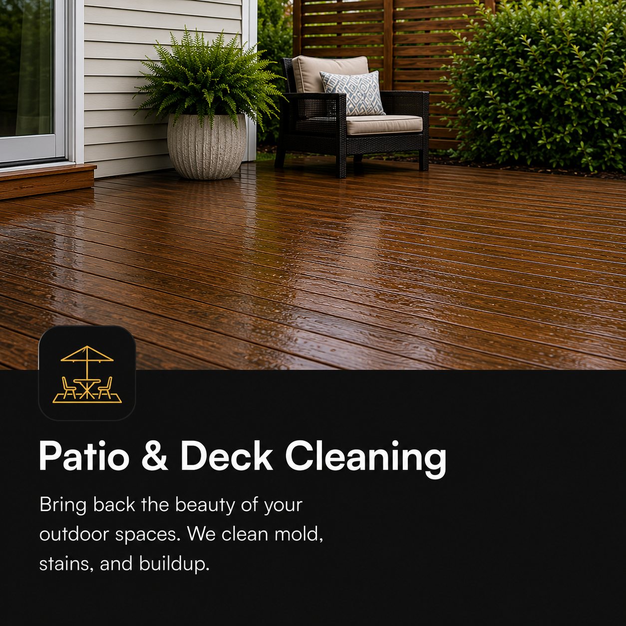 A wet wooden patio with a black outdoor chair with beige cushions and a decorative pillow, a large potted fern, and a wooden privacy screen in the background.