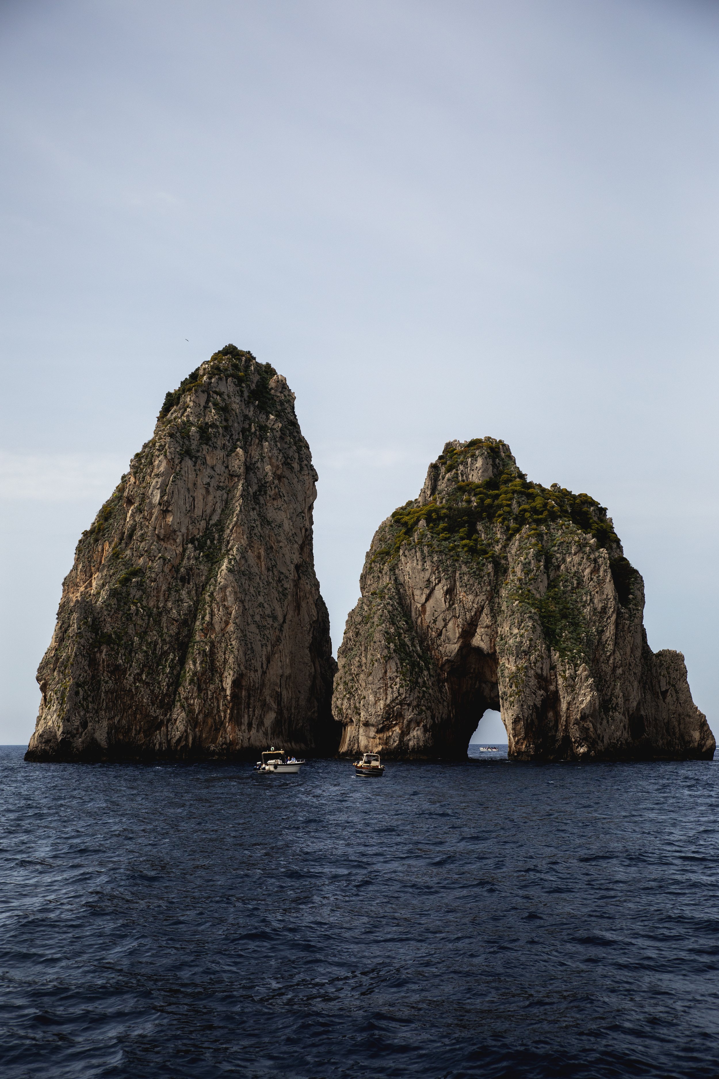 Two large rock formations with natural arch openings in the sea, boats floating nearby, under a cloudy sky.