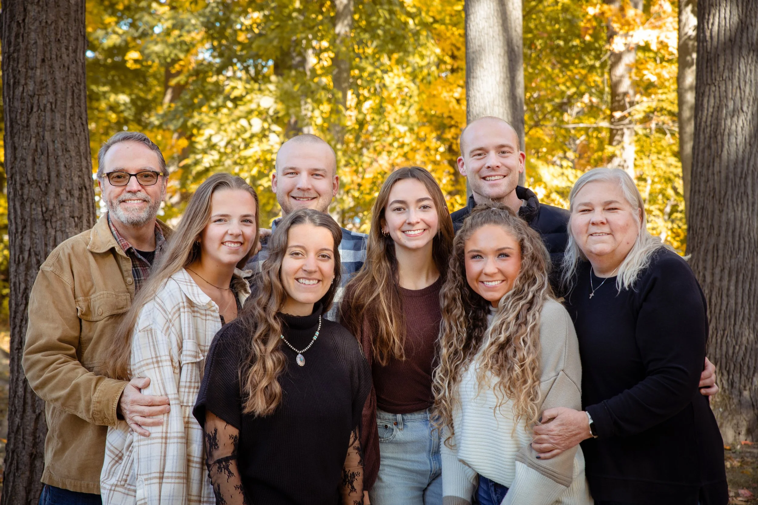 A group of twelve diverse people smiling outdoors during fall, standing among trees with yellow autumn leaves.