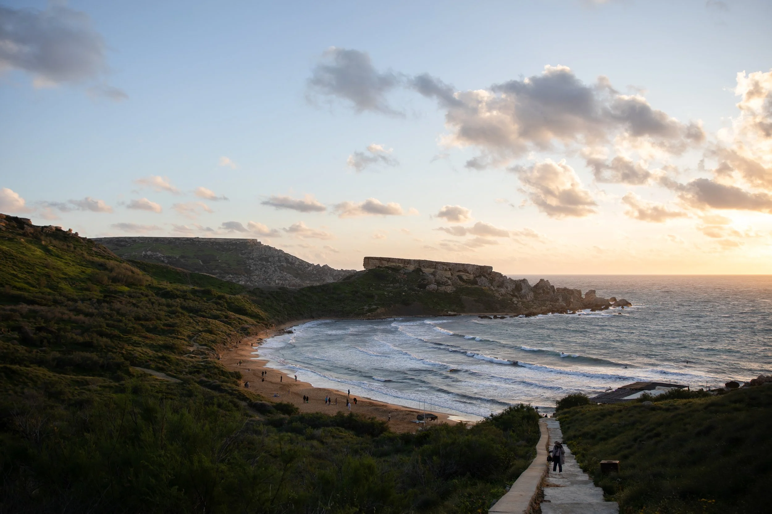 Scenic view of a beach with a pathway leading down from green hills to the sandy shore, with waves crashing on the beach and a sunset in the background.