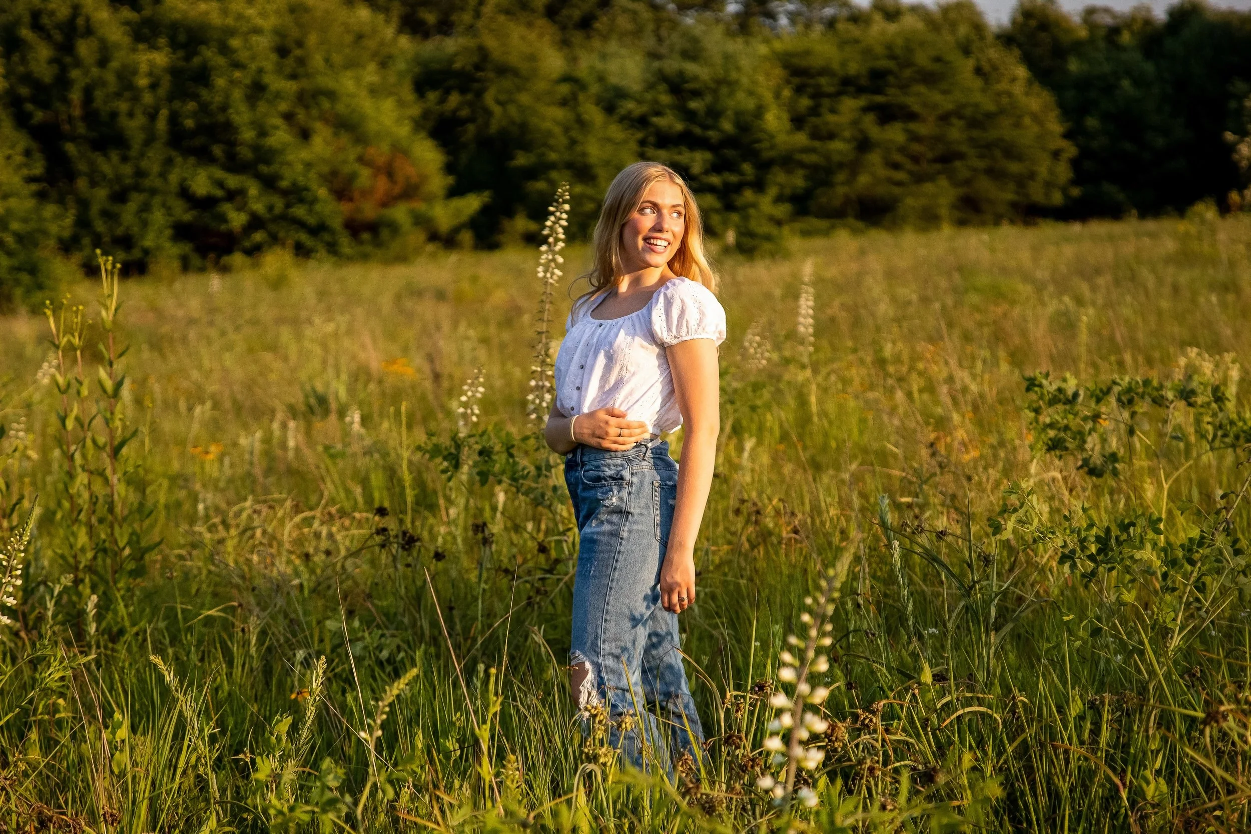 A young woman with blonde hair wearing a white blouse and blue jeans standing in a grassy field during sunset.