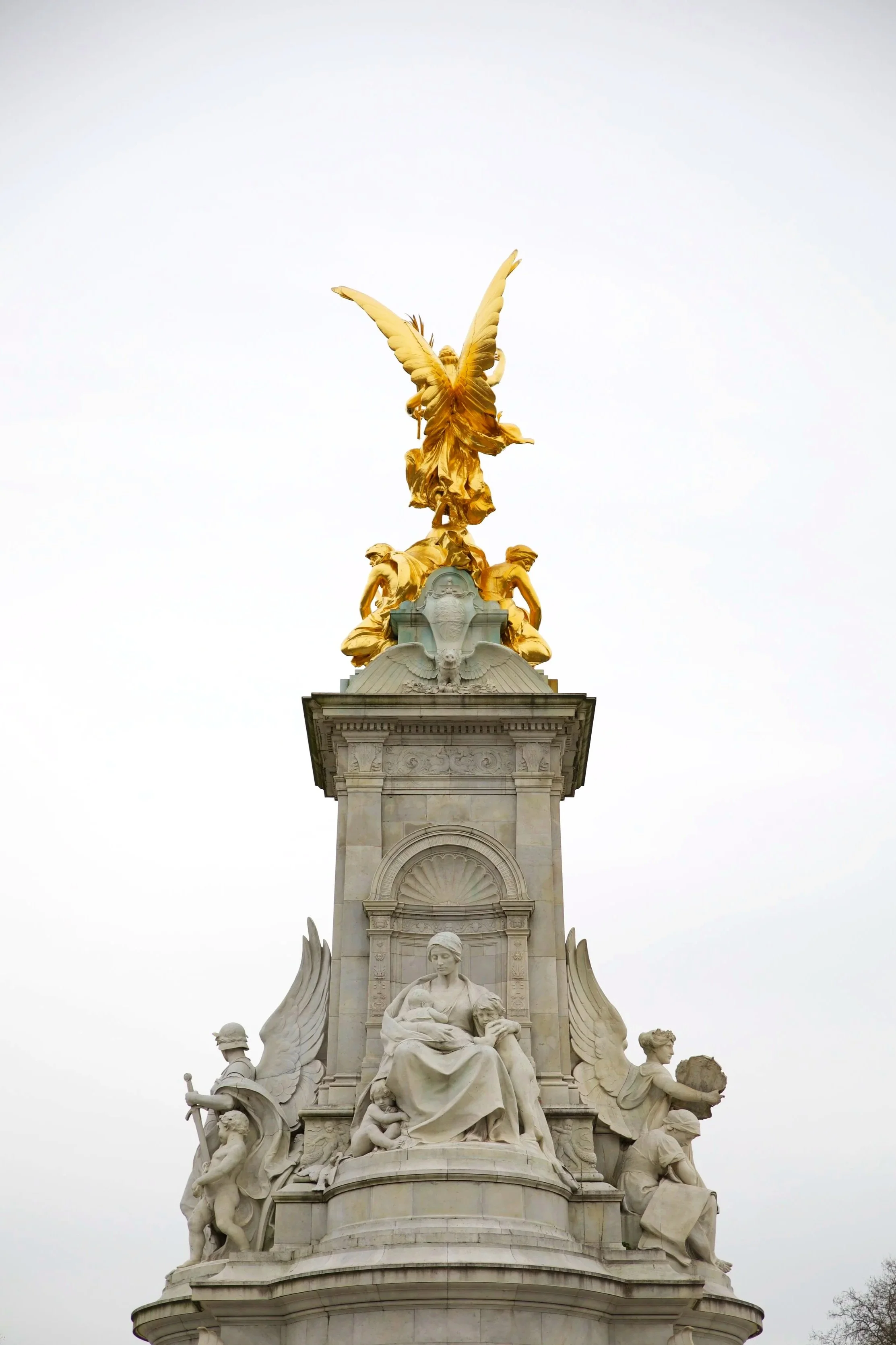 A tall monument topped with a golden sculpture of a winged figure holding a wreath. The monument features multiple white statues and decorative elements on its base, set against a cloudy sky.