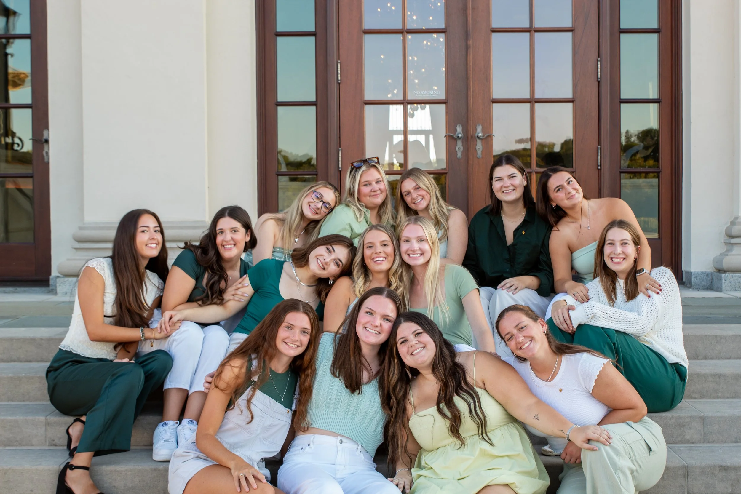 A group of young women sitting on the steps in front of a building with wooden doors, smiling and posing together.
