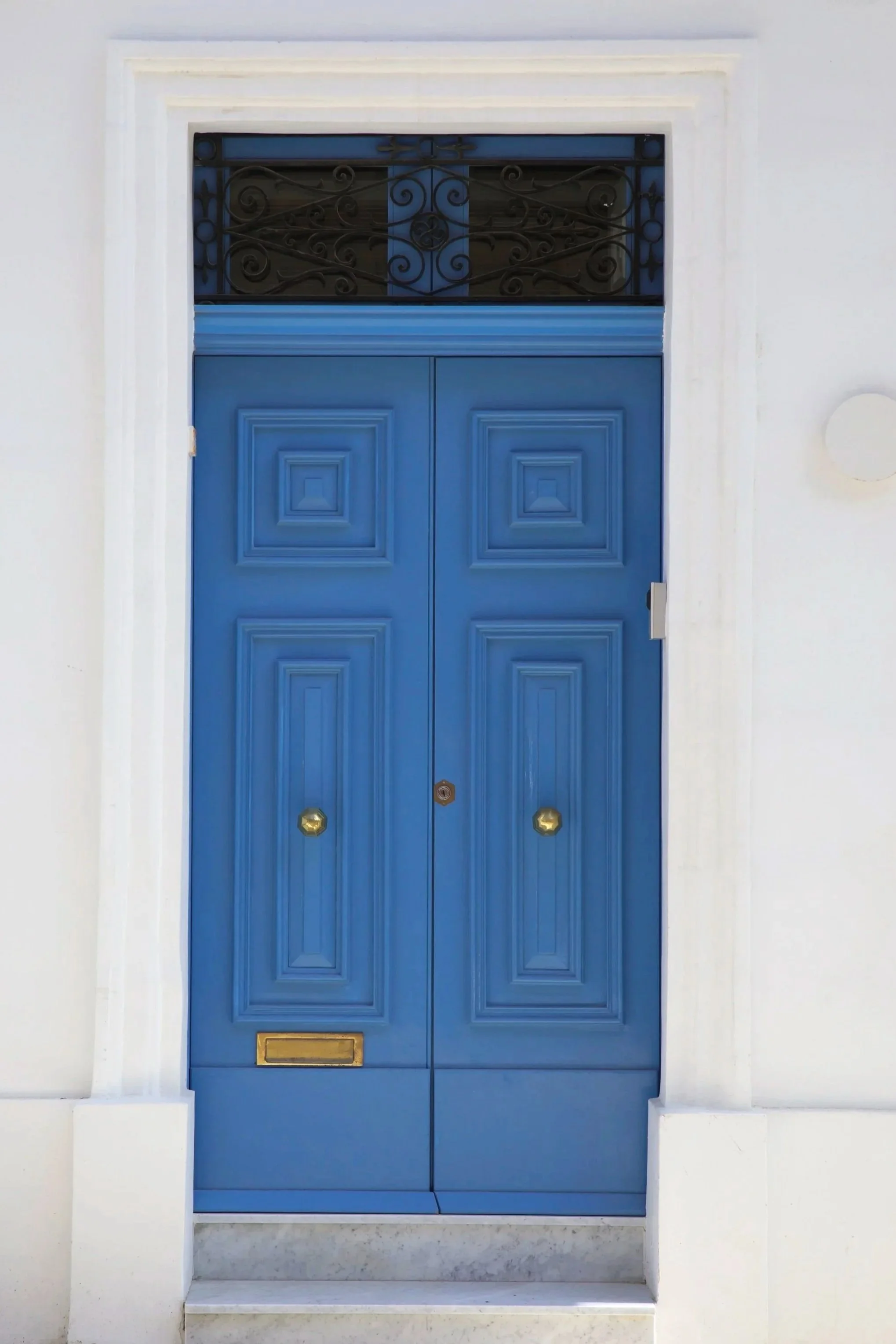 Blue front door with four panel design, brass door handles, mail slot, and a transom window with decorative black iron grill above, set in a white wall.