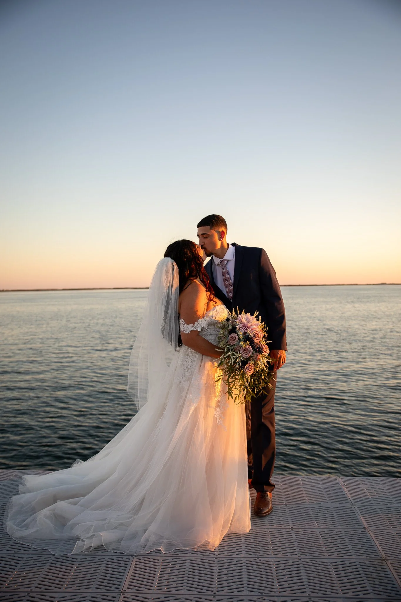 A bride and groom sharing a kiss at sunset by the water. The bride wears a white wedding gown with a long train and veil, holding a bouquet of pink and white flowers. The groom wears a dark suit with a patterned tie.