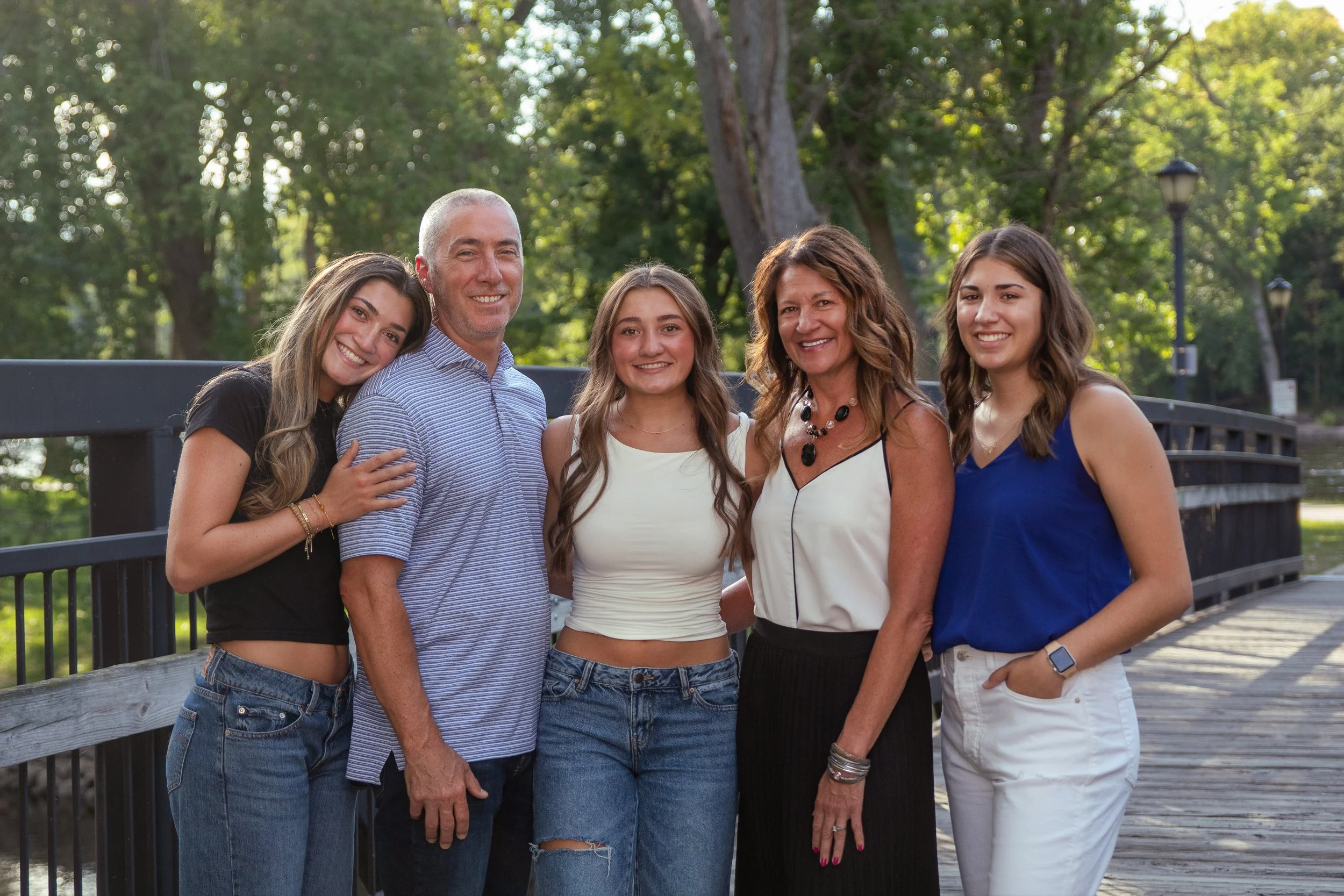 Group of five people smiling outdoors near a bridge with green trees in the background.