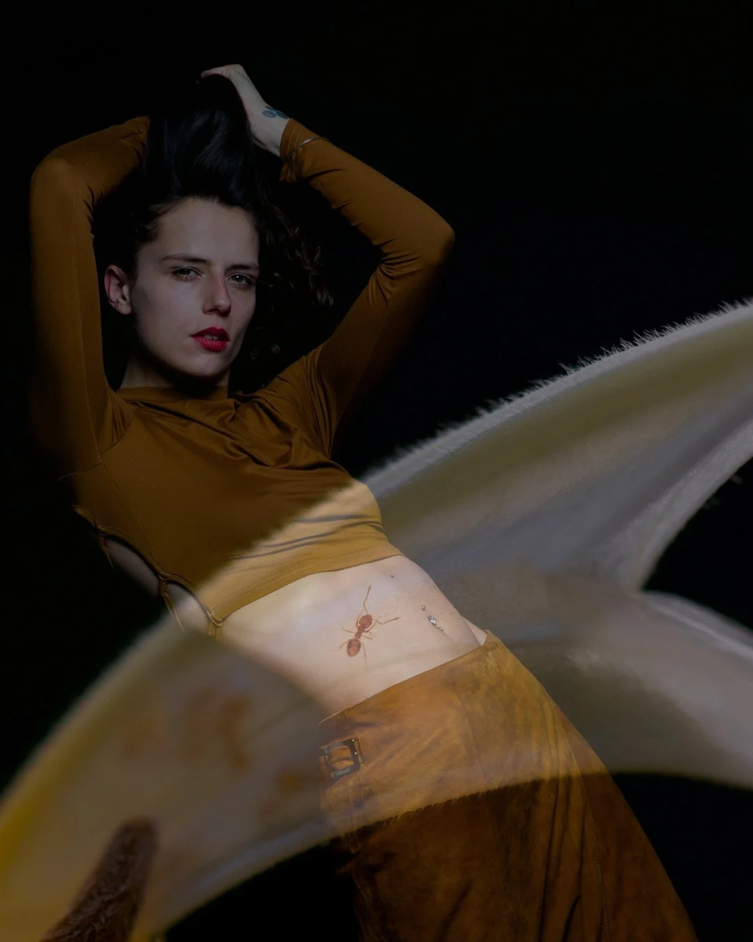 A woman wearing a brown outfit, lying on her back with her hands in her hair, with a reflection of an ant and some jewelry on her stomach, against a black background.