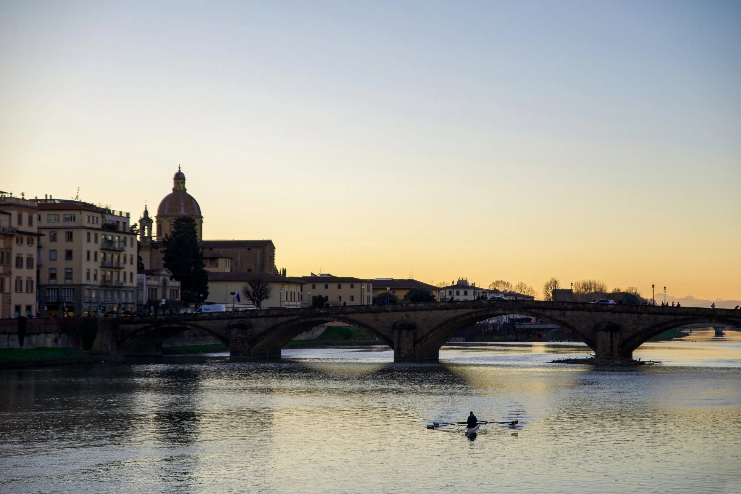 A river scene at sunset with a person rowing a boat in the water, historic buildings with domed rooftops and a bridge crossing the river, and a clear sky with faint light from the setting sun.