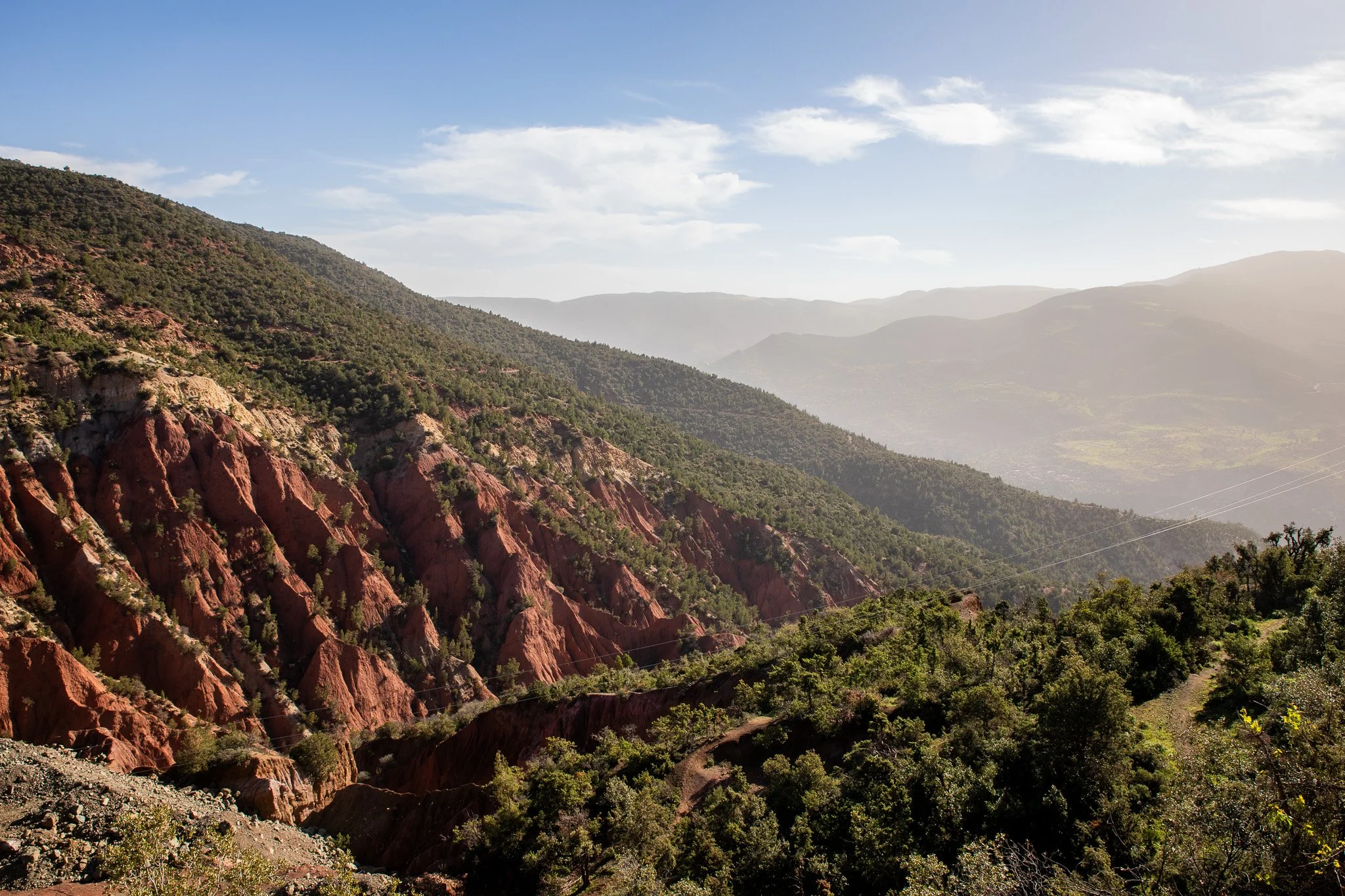 Scenic view of a mountainous landscape with green vegetation, reddish rocky slopes, and distant mountain ranges under a partly cloudy sky.