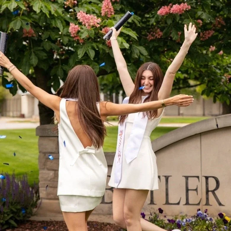 Two women celebrating outdoors with confetti, one with a graduation sash, near a sign that says "Fletcher".