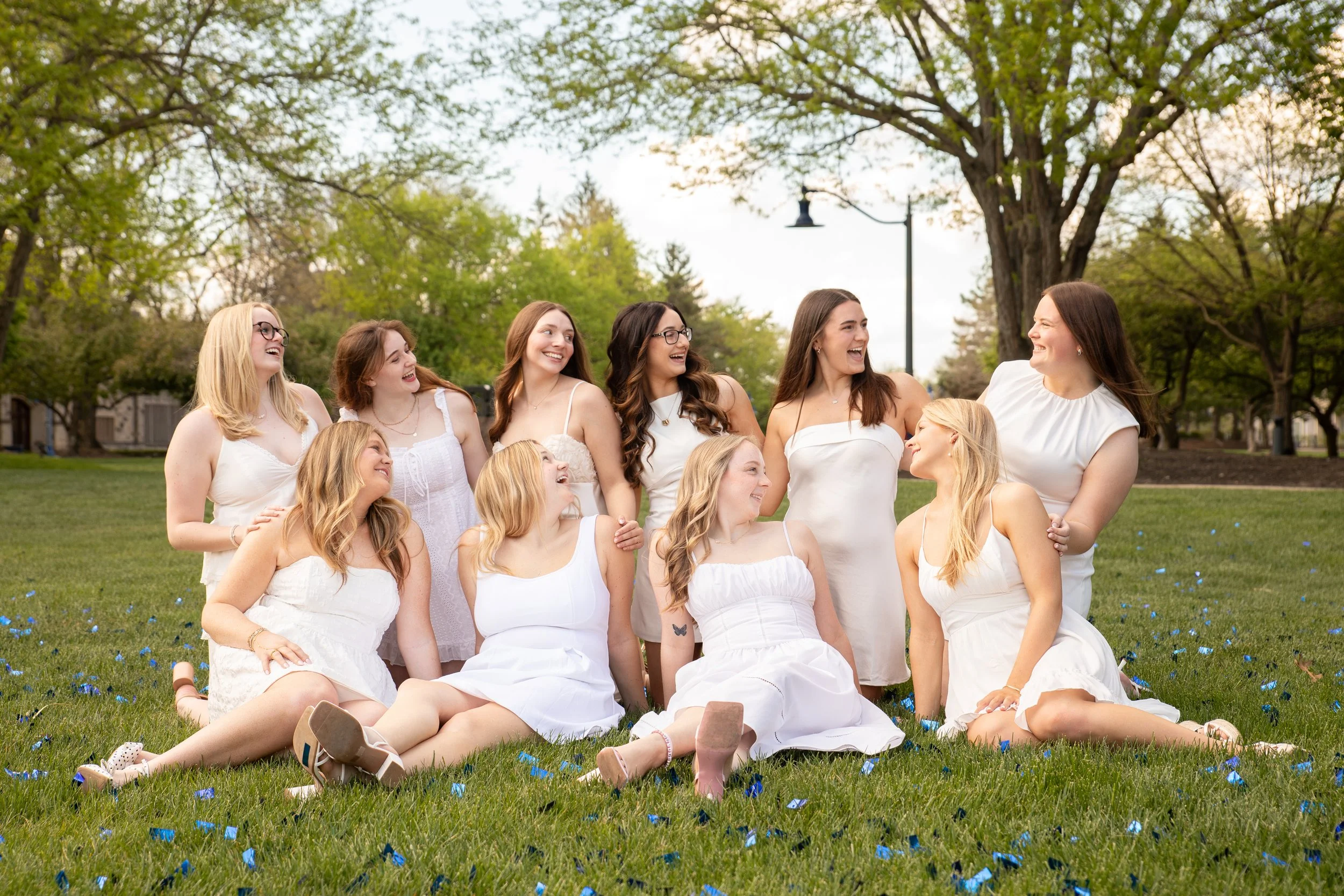A group of women and girls dressed in white, smiling and laughing together in a park with green trees and a large tree in the background.