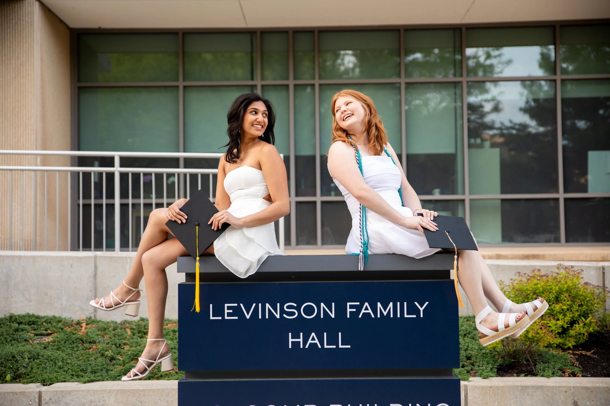 Two women in white dresses sitting on a platform holding diplomas, smiling and looking at each other, in front of a building with large glass windows. A sign below them reads "LEVINSO FAMILY HALL."