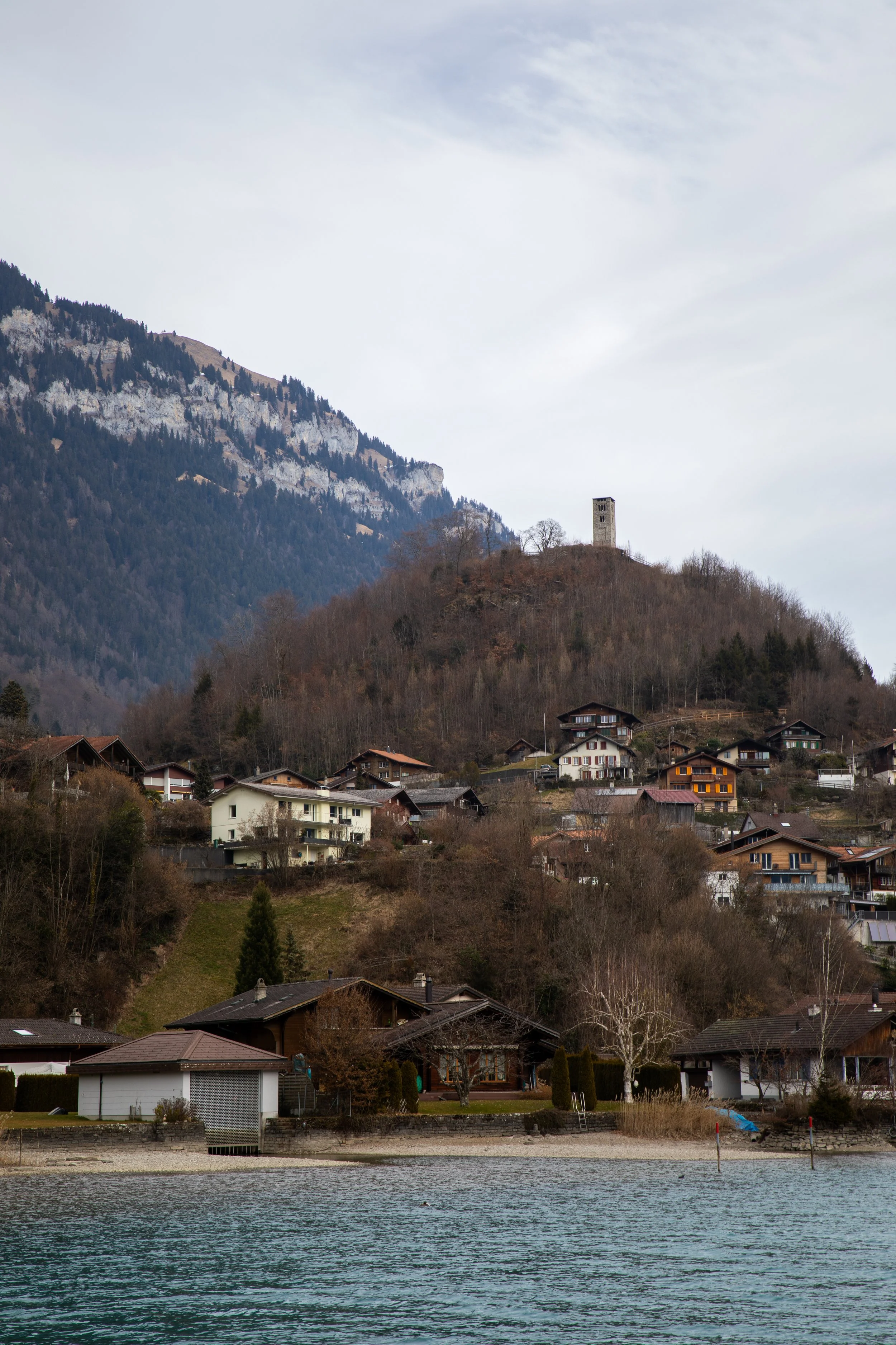 A lakeside village at the base of a mountainous hillside, with houses and trees in the foreground and a castle or tower on the hilltop, under a cloudy sky.