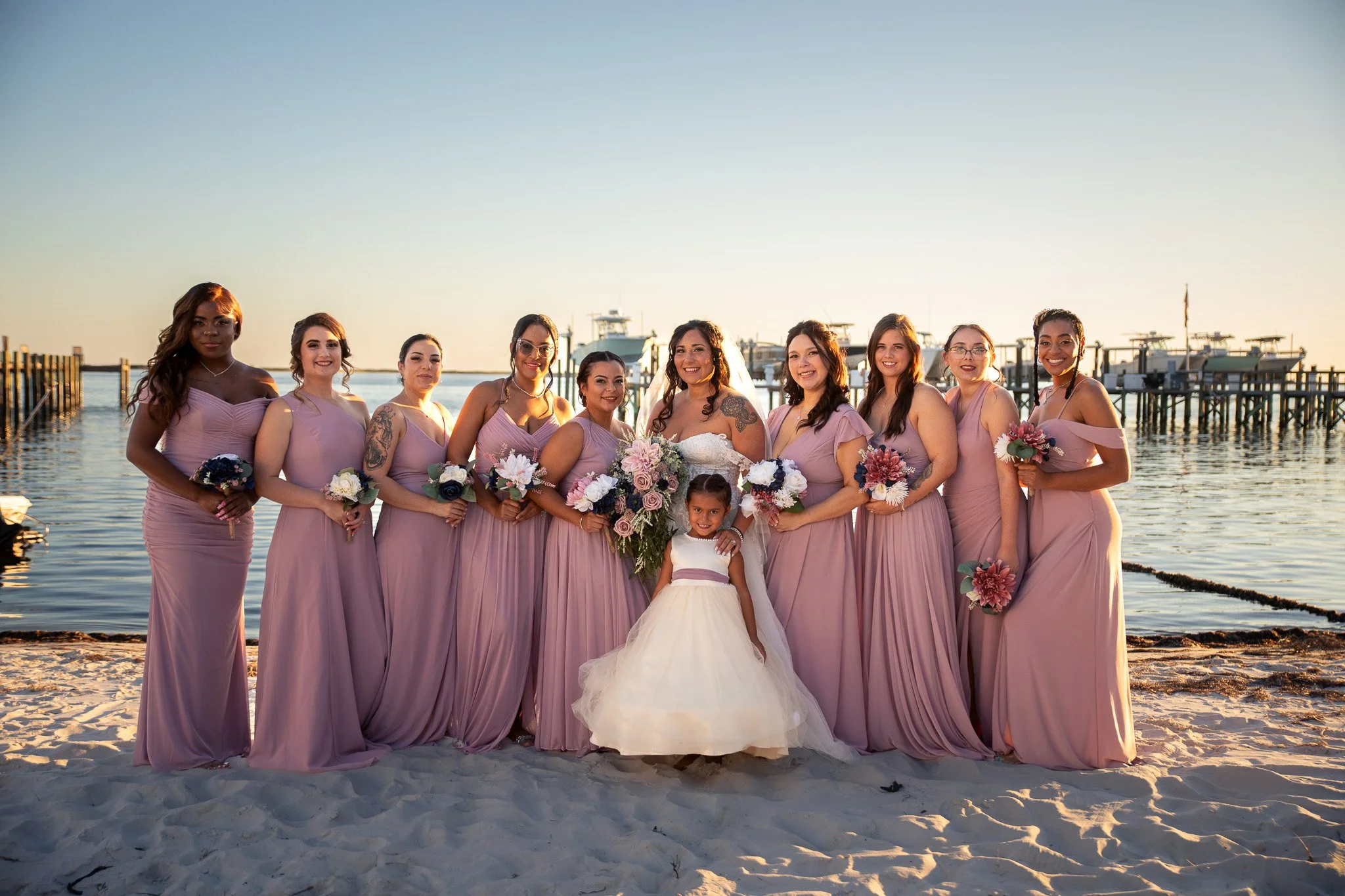A group of women and girls in pink dresses, standing on a sandy beach near the water, with boats and a pier in the background, during sunset.