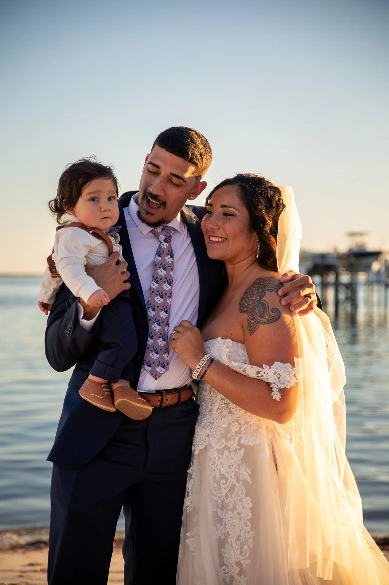 A couple dressed in wedding attire stands closely with their arms around each other at the beach during sunset, smiling and enjoying the moment. The groom is holding a small child in his arm.