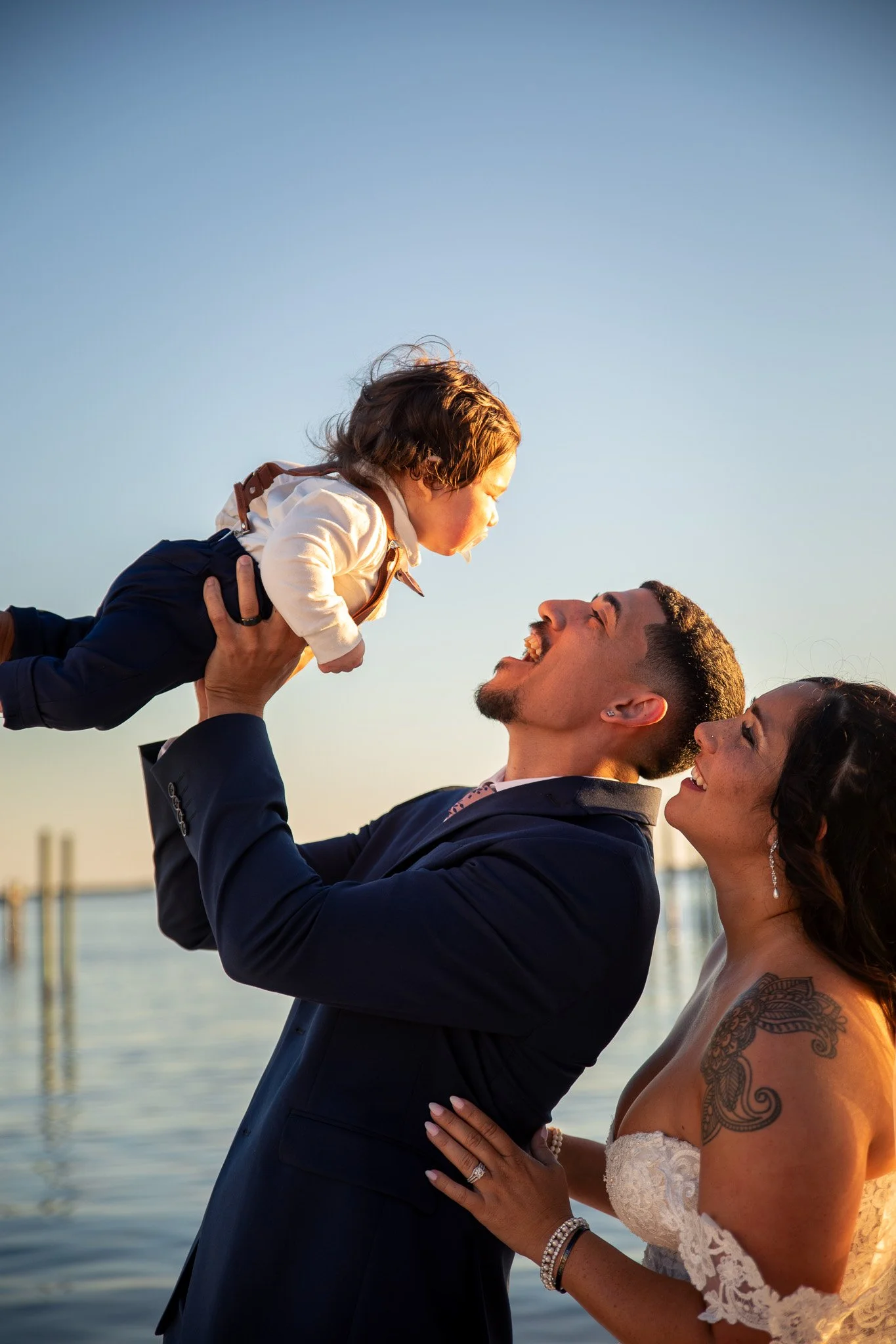 A groom lifts a young girl in a white jacket and dark pants, blowing kisses in a heartfelt moment during a wedding ceremony outdoors near a body of water.