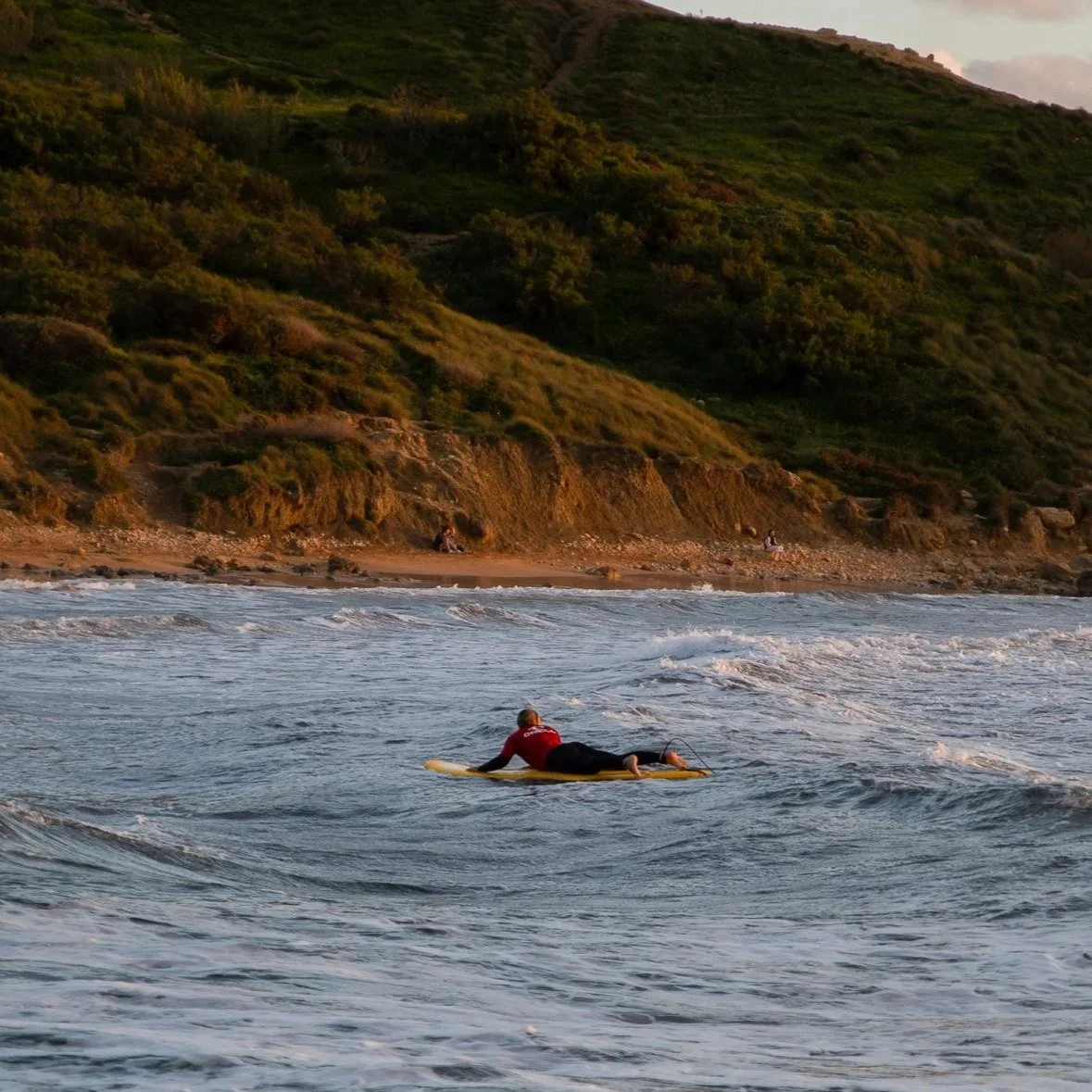 A person in a red shirt surfing on a yellow surfboard in the ocean near a hilly coastline with green vegetation at sunset.