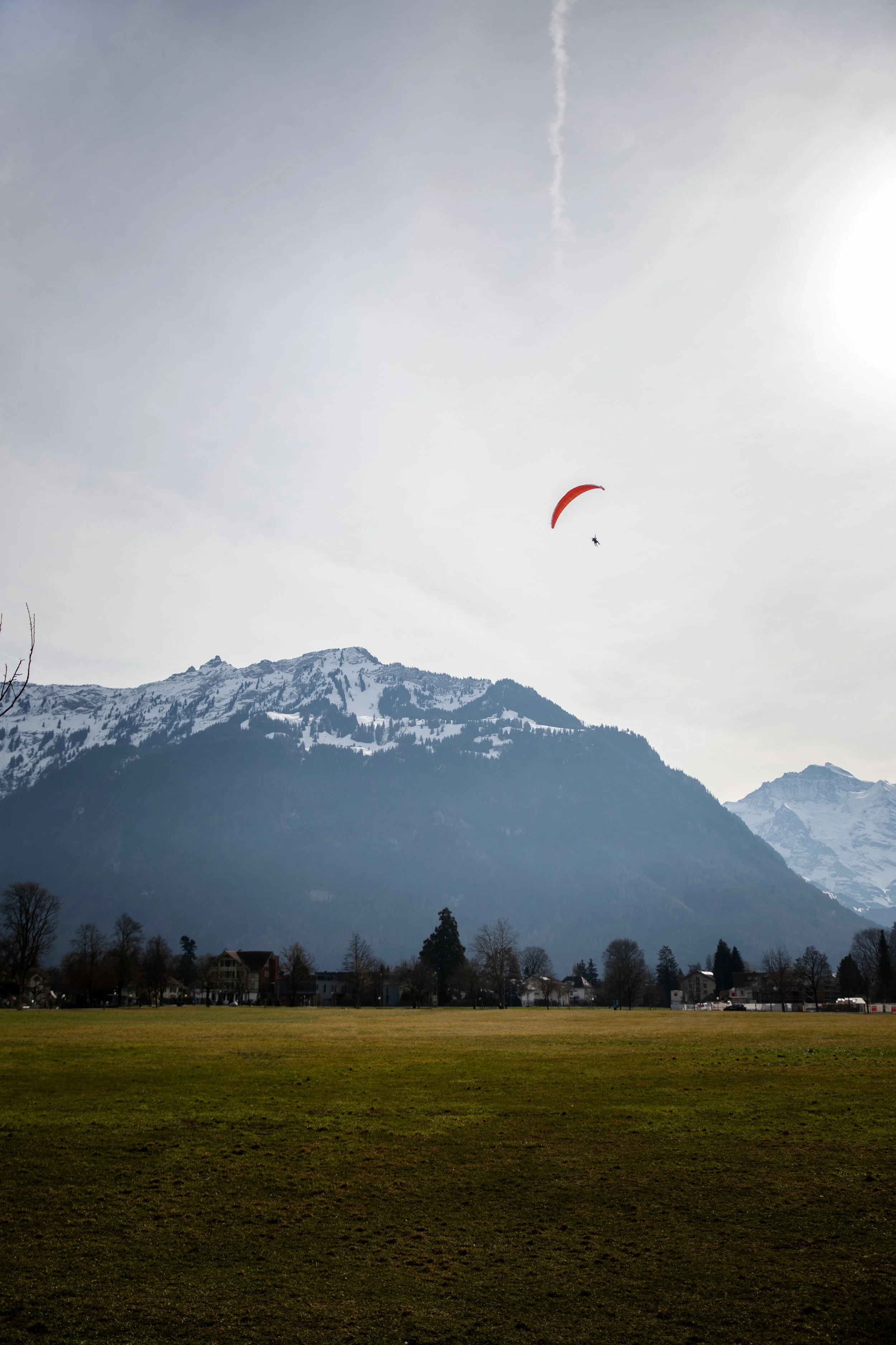 A scenic landscape with a mountain range, snow-capped peaks, and a grassy field in the foreground. There is a paraglider with an orange parachute flying in the sky. Houses are visible at the base of the mountains.