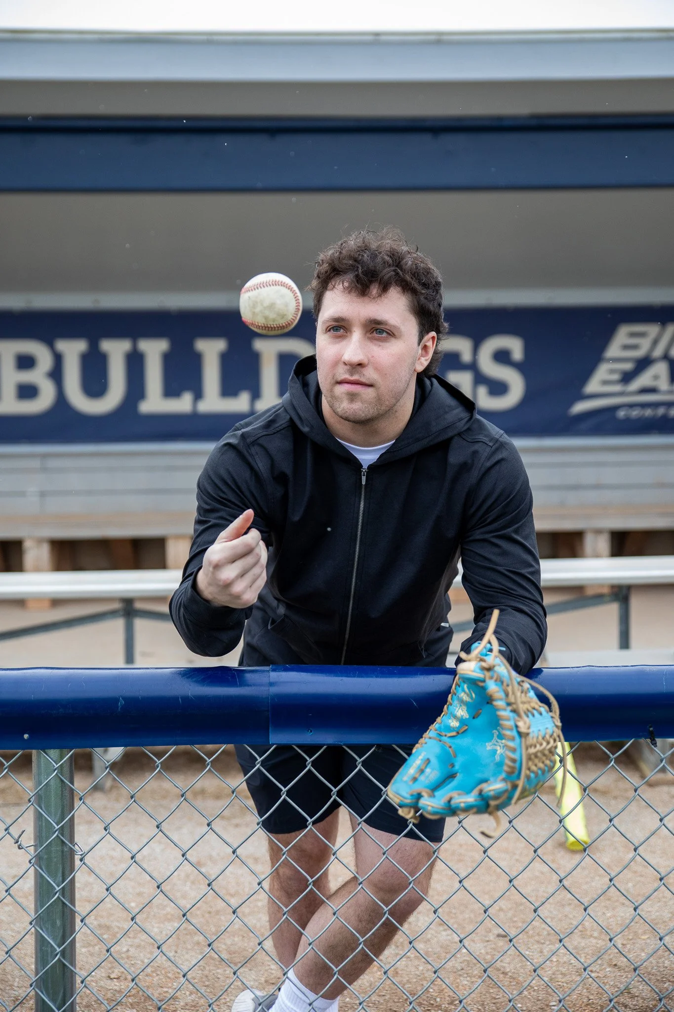A young man in athletic clothing with curly hair is sitting on a bench, ready to catch a baseball. There is a baseball glove placed on a metal railing in front of him. The background shows a sign with the word 'BULLDOGS'.