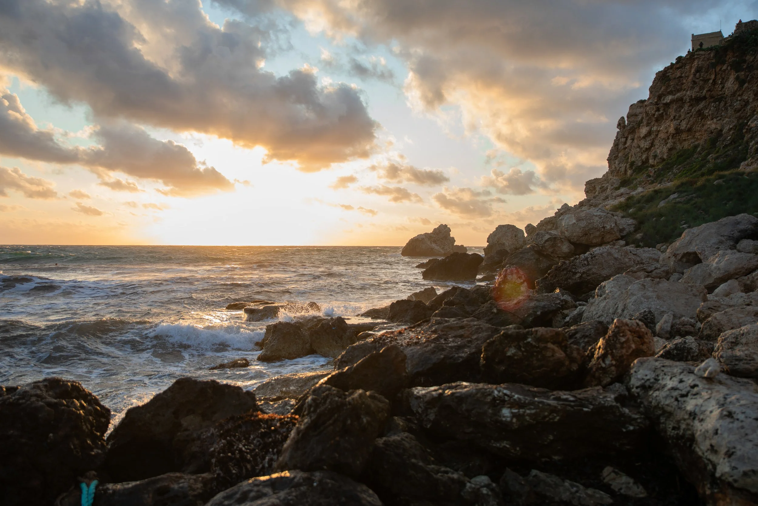 Sunset over the ocean with clouds and a rocky shoreline in the foreground.