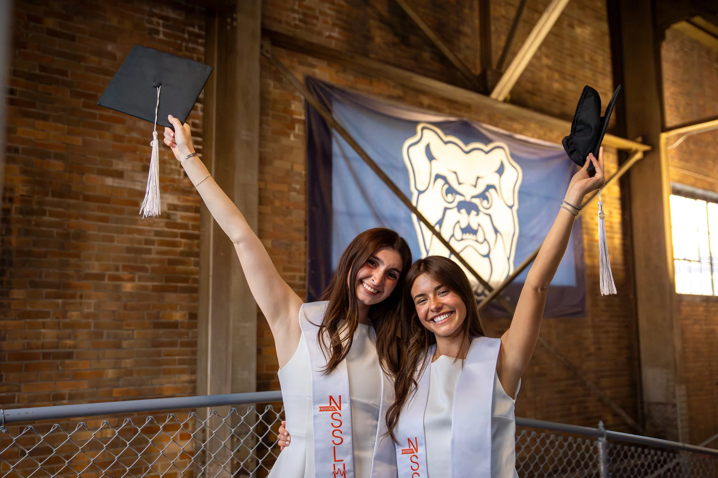 Two young women in white dresses celebrating graduation, holding mortarboards in the air, standing in front of a chain-link fence with a brick wall and a large Bulldogs banner in the background.