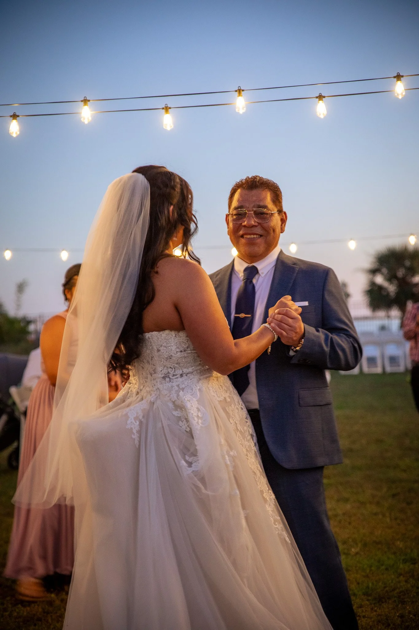 A bride and groom dancing at an outdoor wedding reception during evening with string lights overhead.