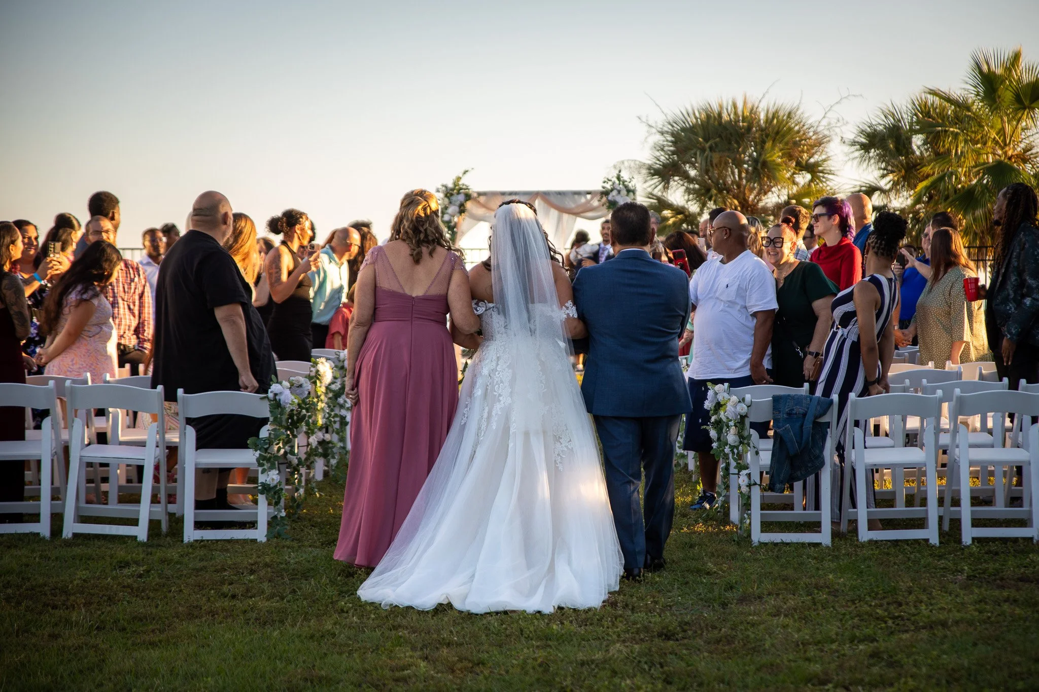 Outdoor wedding ceremony with bride and guests. Bride in white wedding dress and veil, flanked by bridesmaid in pink dress and groom in blue suit, walking towards altar with floral arrangements, as guests watch and take photos. Palm trees and clear sky in background.
