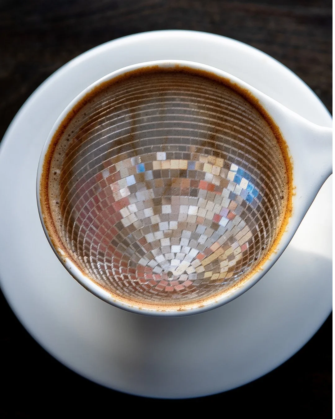 Top view of a cup of coffee with a mirror-like mosaic pattern on the inside, placed on a small white saucer.