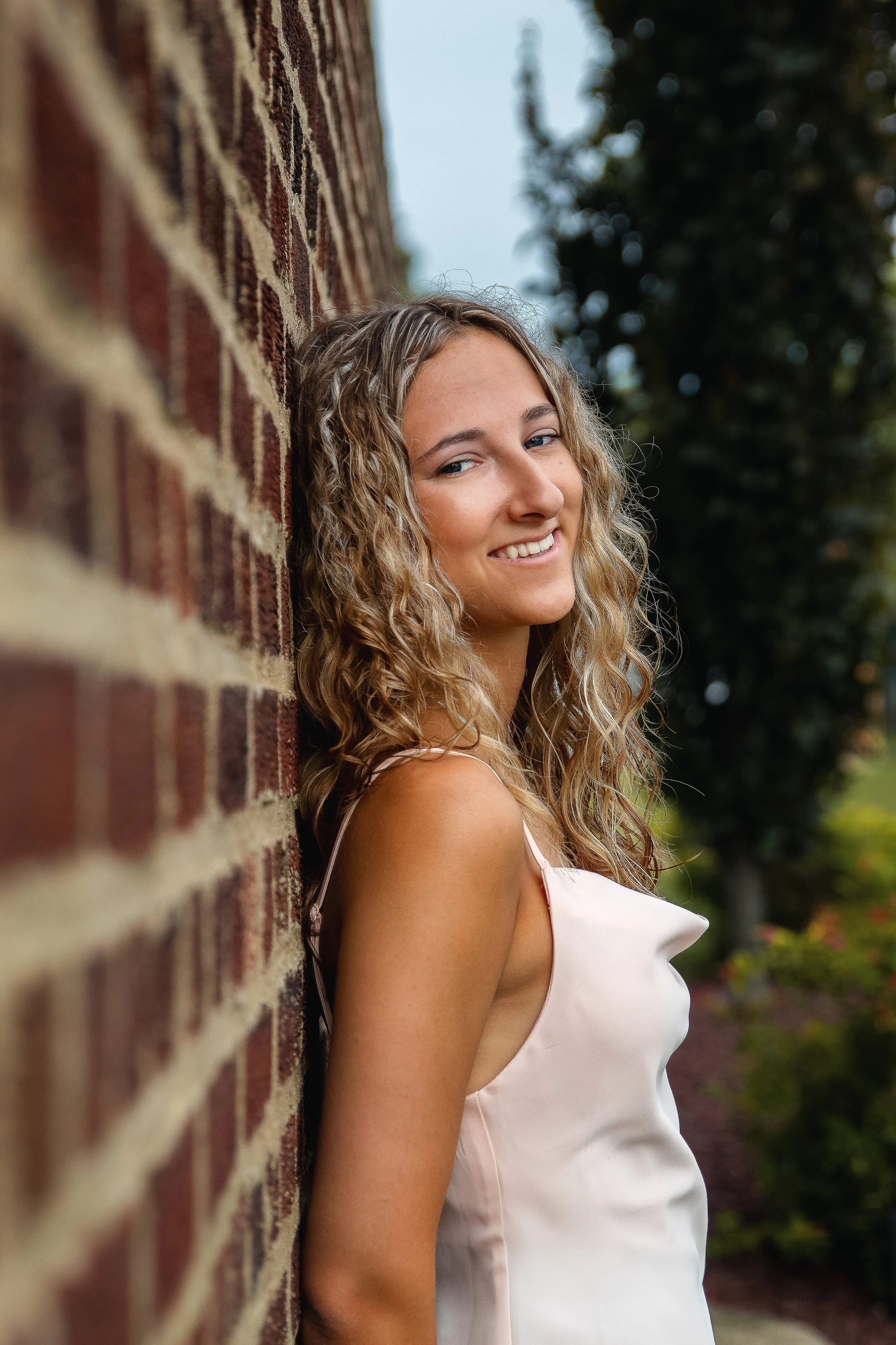 A young woman with blond, curly hair smiling and leaning against a brick wall outdoors, with greenery and trees in the background.
