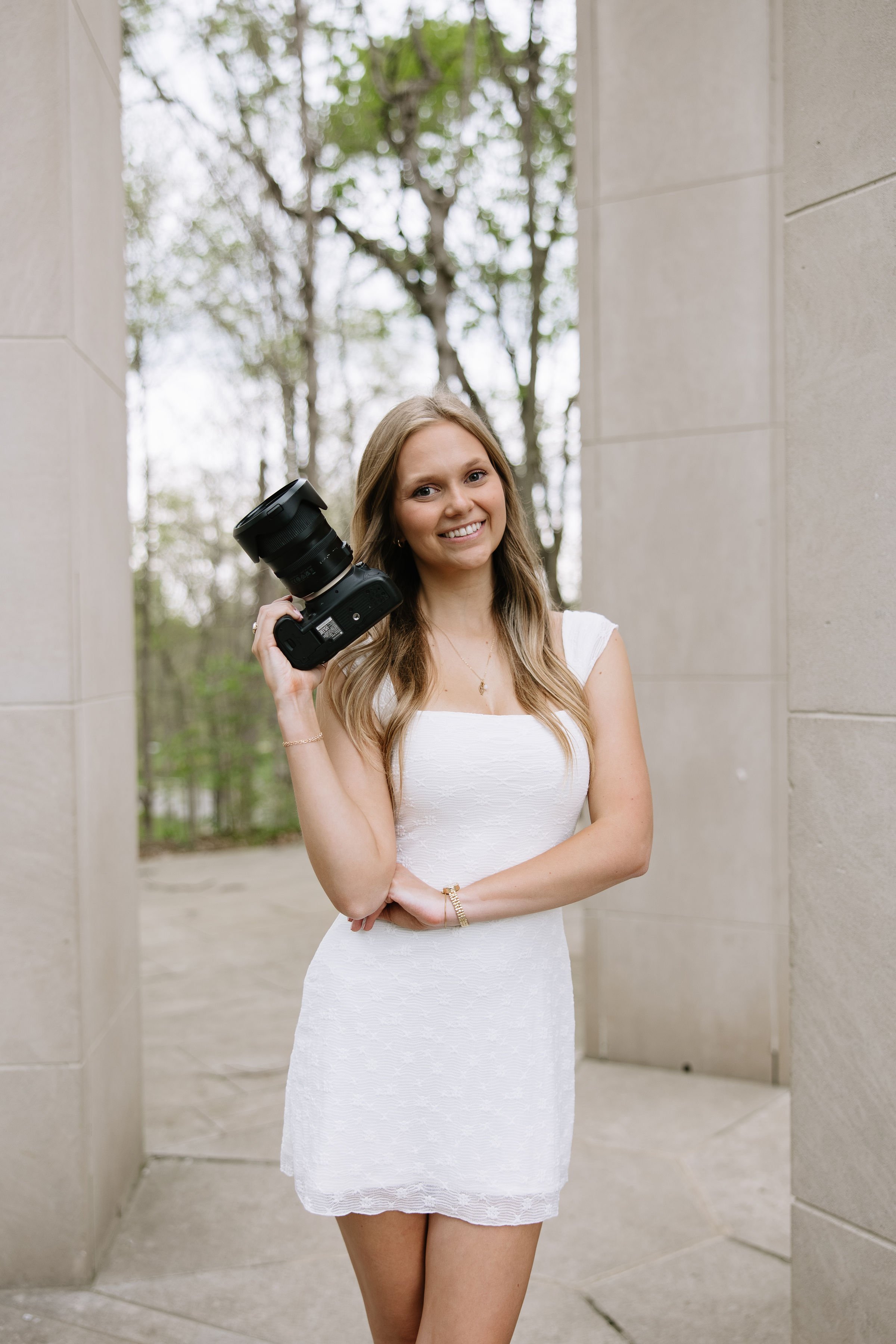 A woman in a white dress holds a camera and smiles at the camera.