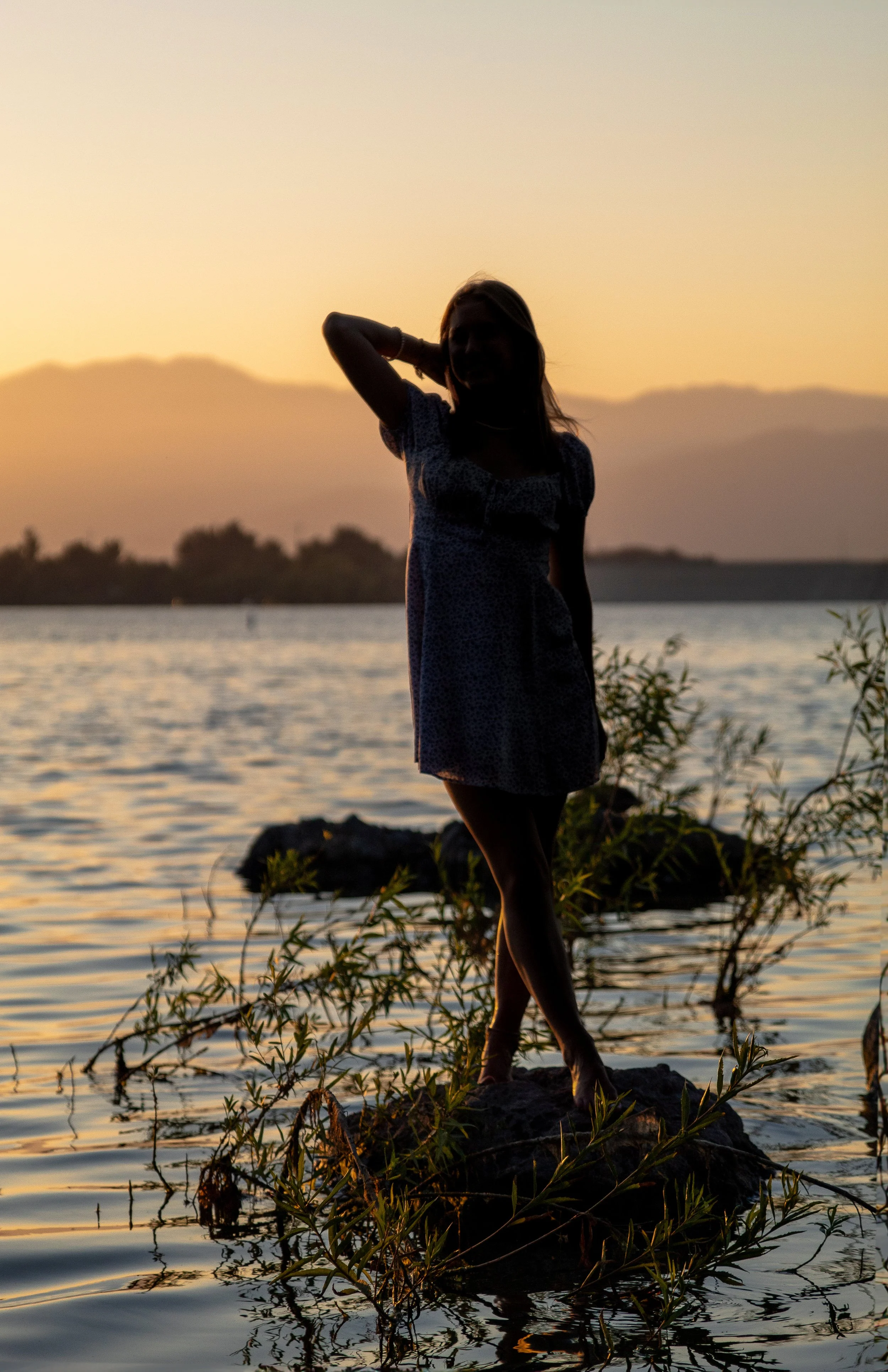 A silhouette of a woman standing on a rock in the water during sunset with mountains in the background.