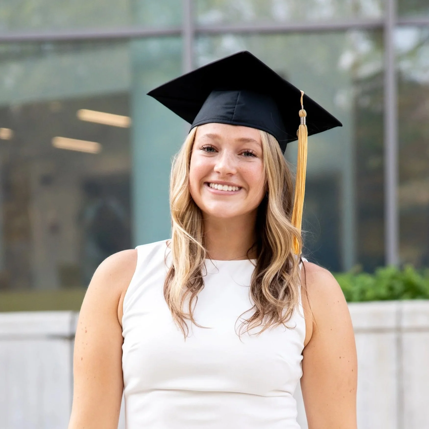 Close-up of a smiling young woman wearing a black graduation cap with a gold tassel, standing outdoors in front of a modern building with large glass windows.