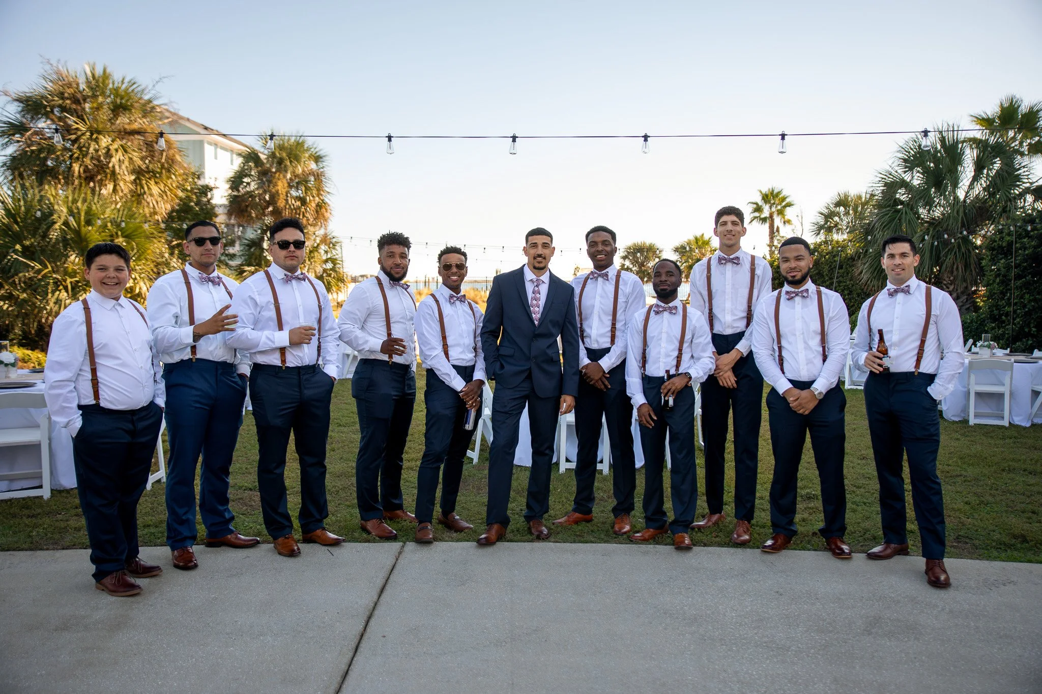 A group of eleven men dressed in formal attire, including white shirts, suspenders, bow ties, and dress shoes, standing outdoors during the day with a background of palm trees and string lights, smiling at the camera.