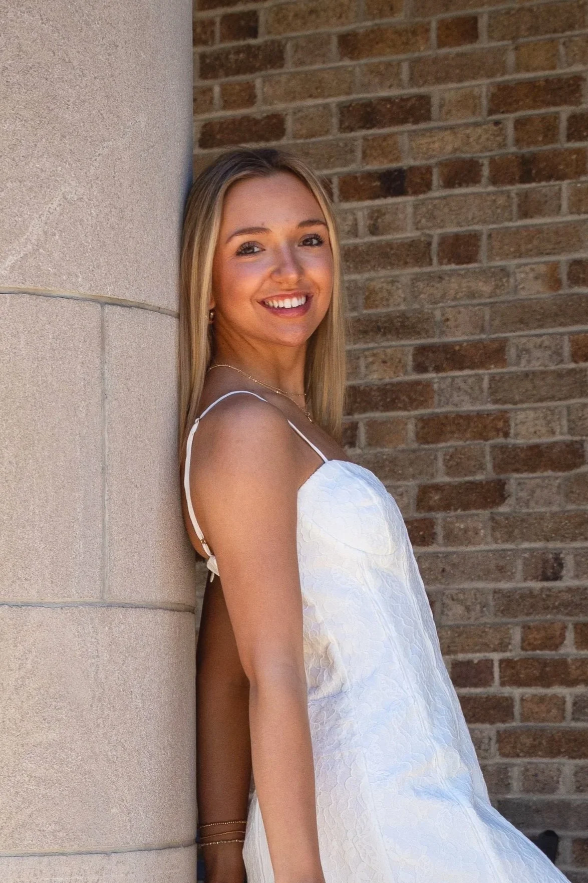 A young woman with blonde hair wearing a white dress with thin straps stands outdoors, leaning against a beige stone column with a brick wall in the background, smiling at the camera.