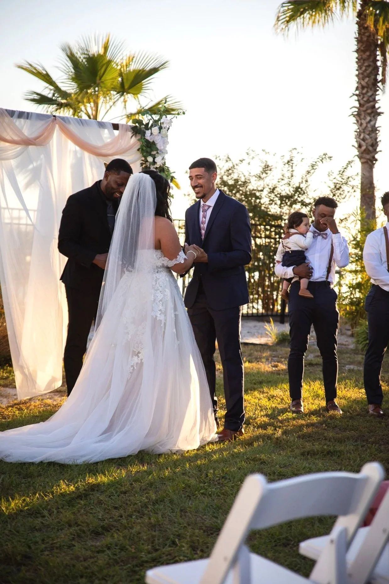 A wedding ceremony outdoors during sunset with couples exchanging vows under a decorated arch, palm trees in the background, and guests seated on white chairs.