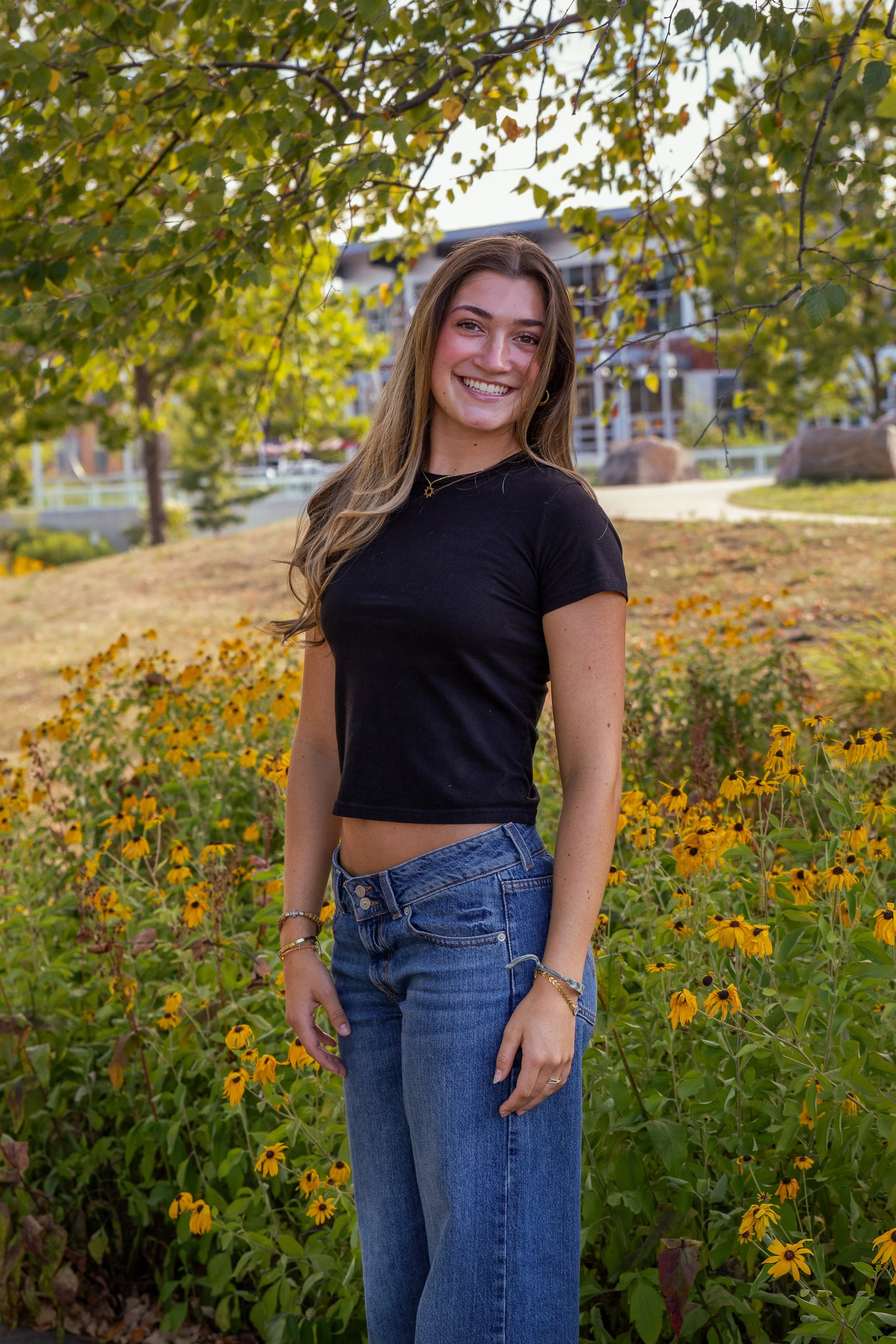 A young woman with long, wavy brown hair smiling while standing outdoors among yellow flowers and green trees in a park during daytime.