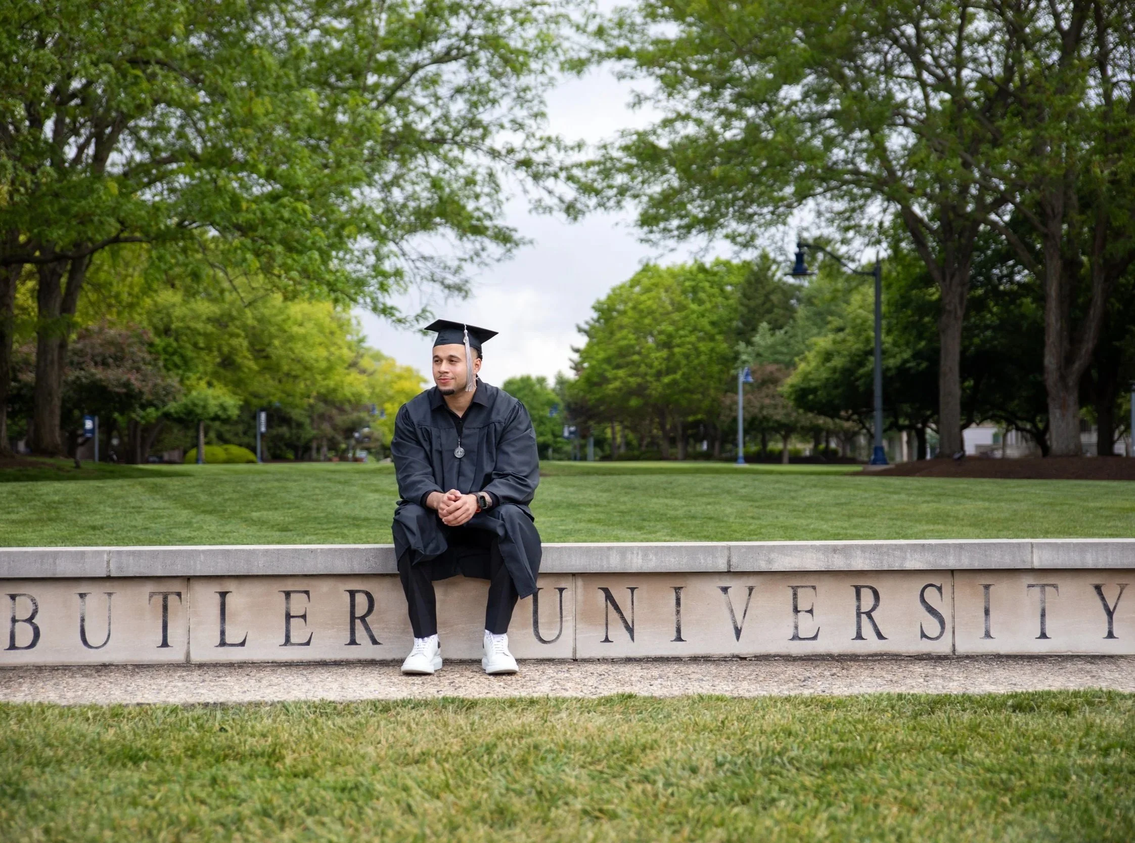 A young man in a graduation gown and cap sitting on a stone ledge in front of a university sign, surrounded by green trees.