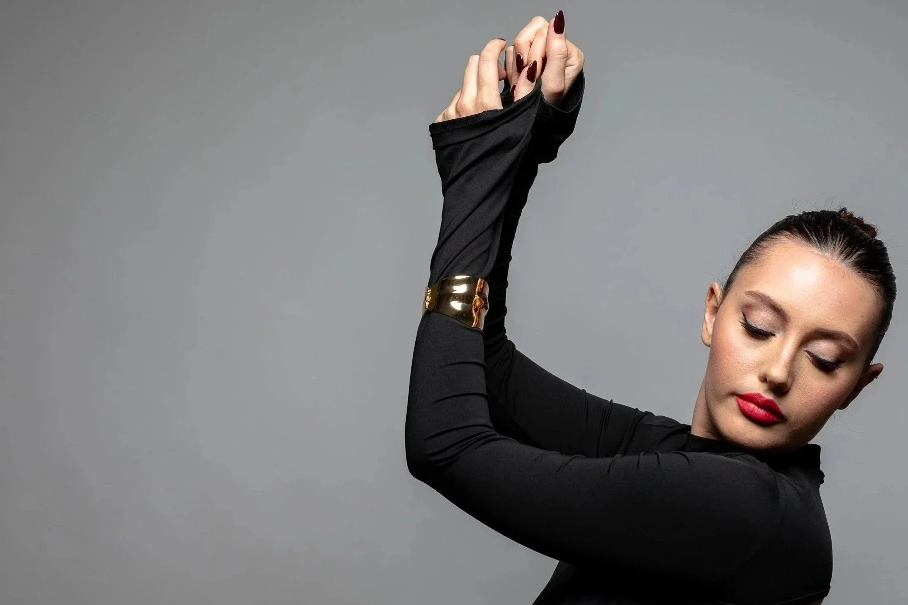 A woman with dark hair and red lipstick poses with her eyes closed, wearing a black long-sleeved top, a metallic bracelet, and stretching her arm upward against a gray background.