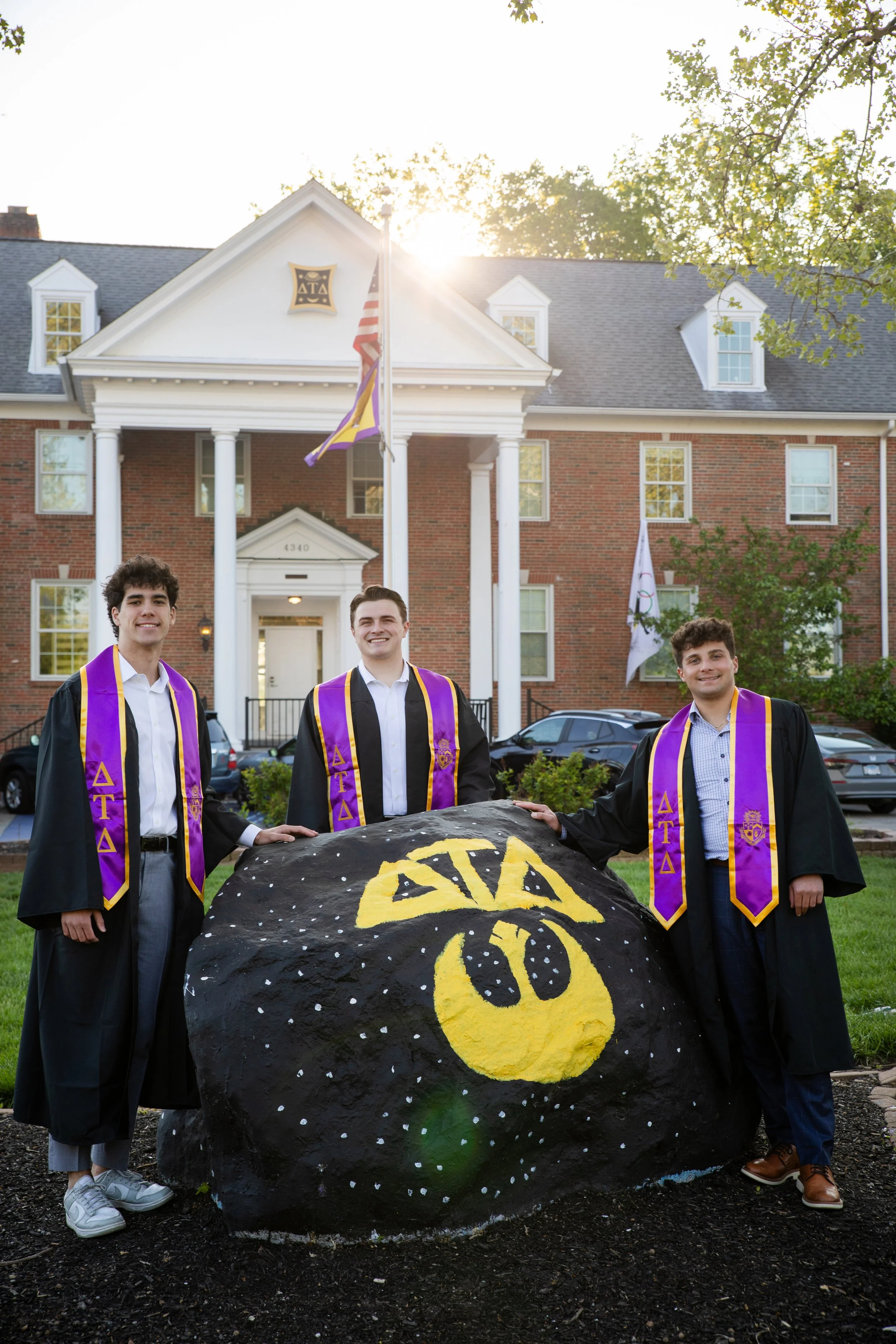 Three young men in graduation gowns and purple stoles with symbols stand outside a brick building with white columns, holding a large painted rock with a yellow Alpha Tau Omega fraternity symbol. The background includes flags, windows, and trees, with sunlight shining above.