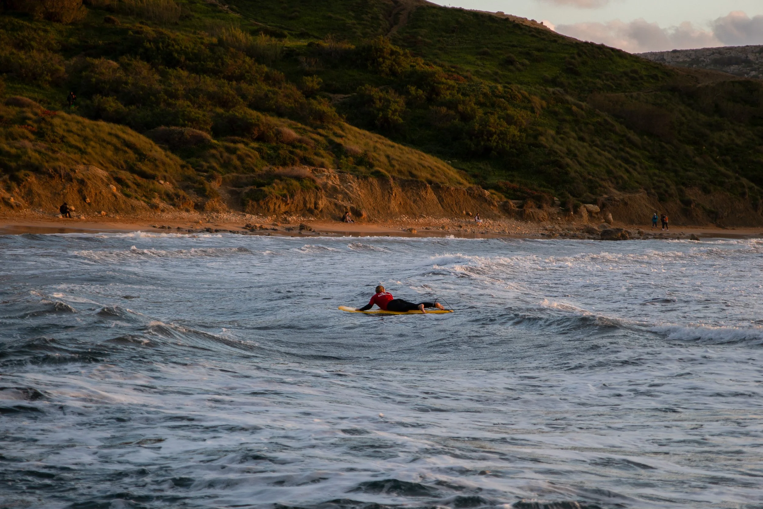 A surfer lying on a yellow surfboard riding a wave in the ocean near a beach with green hills in the background.