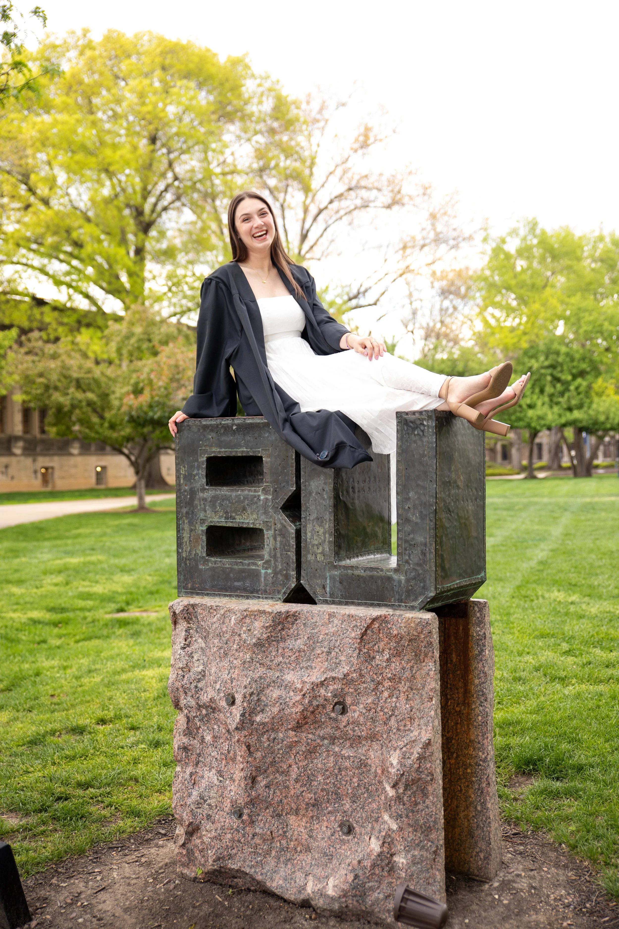 A young woman in graduation attire sitting on a large stone sculpture with smiling face, in a park with green trees in the background.