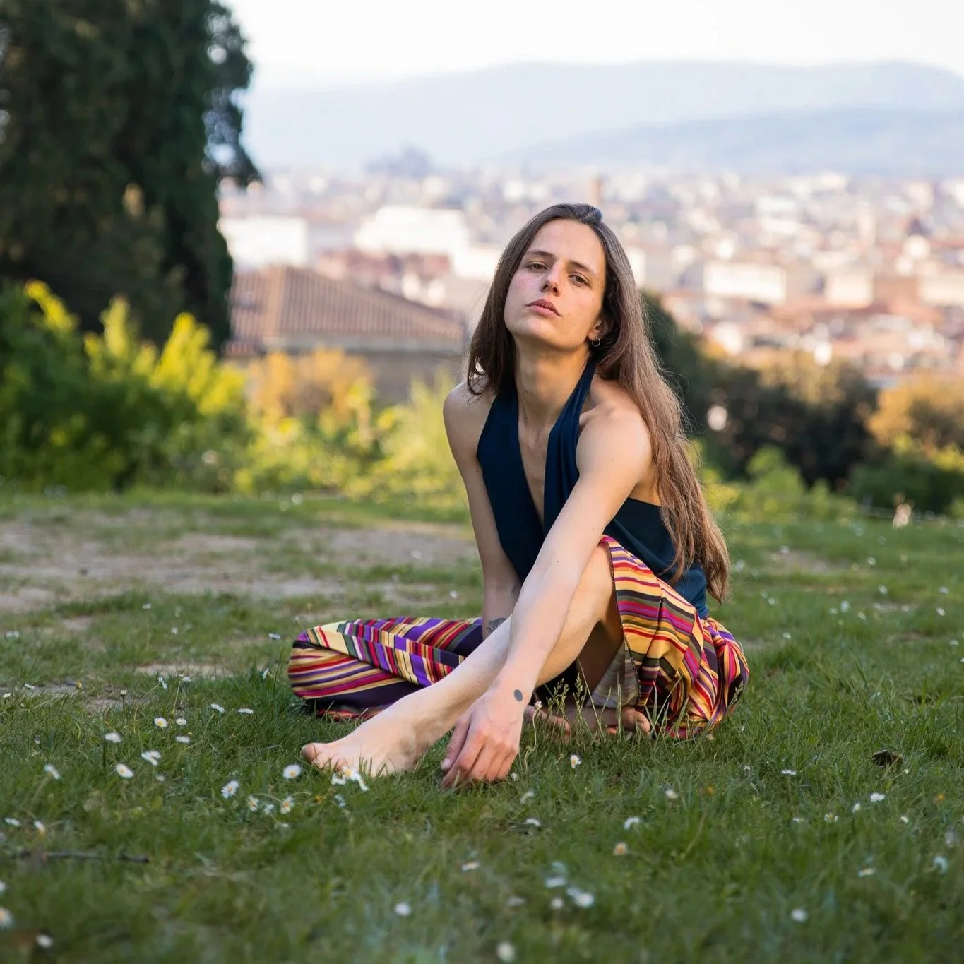 A young woman with long brown hair, wearing a navy blue top and colorful striped pants, sitting on grass in an outdoor park with a cityscape in the background.