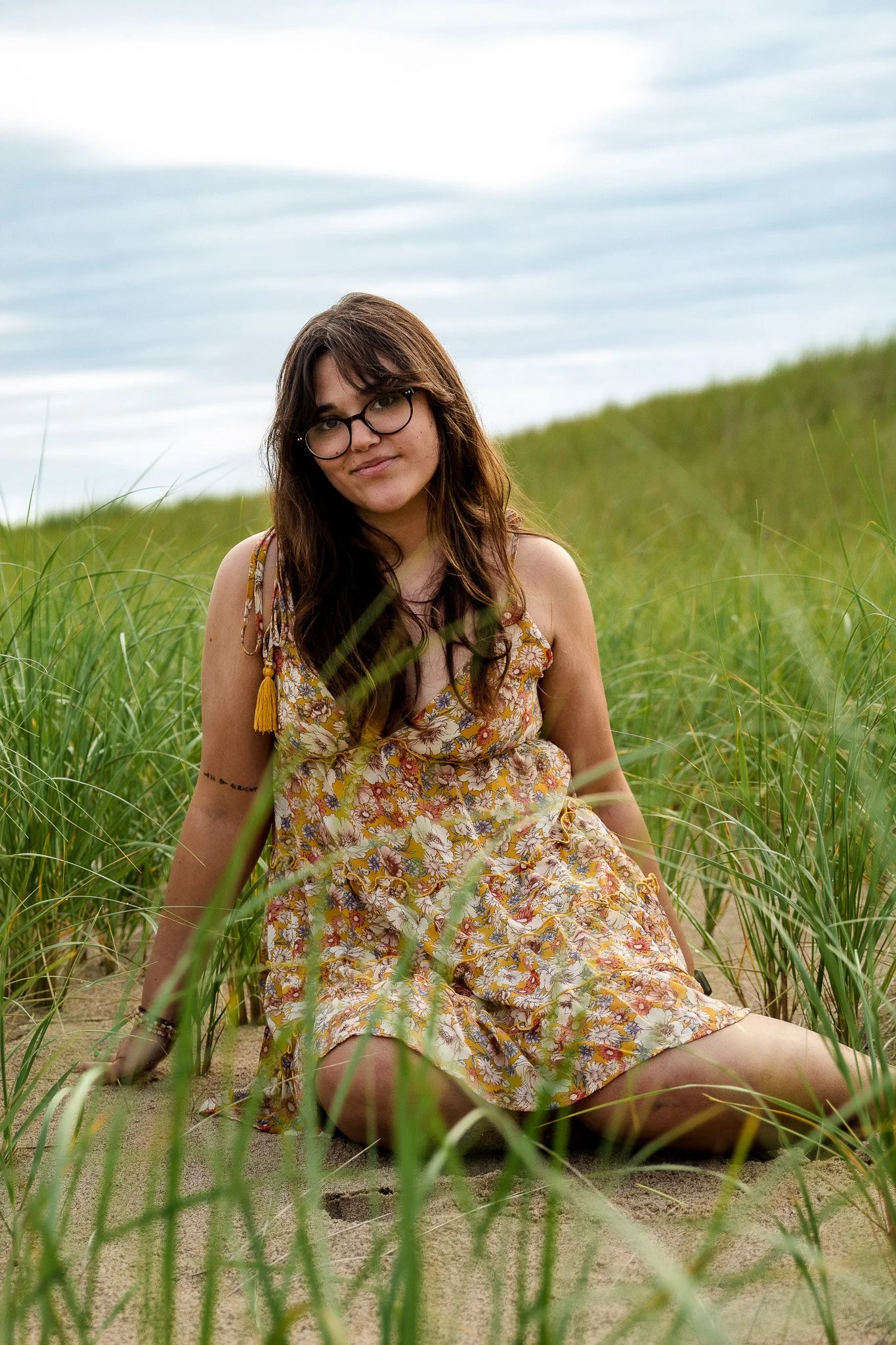 Young woman with glasses sitting in tall grass on a sandy area outdoors, wearing a floral dress, with a cloudy sky in the background.
