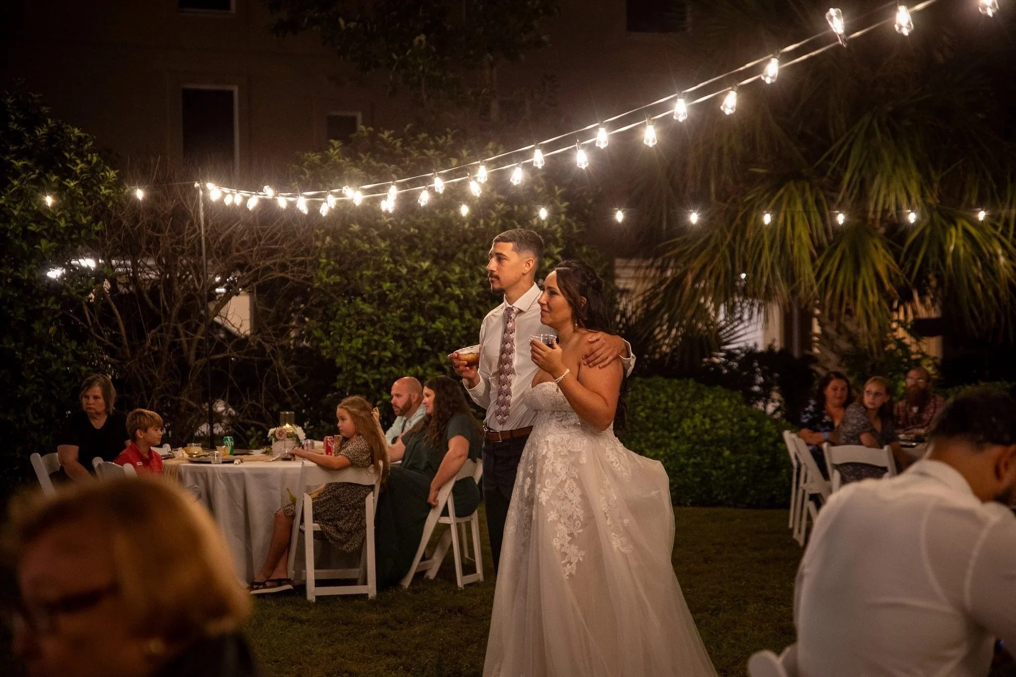 A bride and groom at night, standing close together, surrounded by wedding guests at outdoor reception, with string lights overhead.