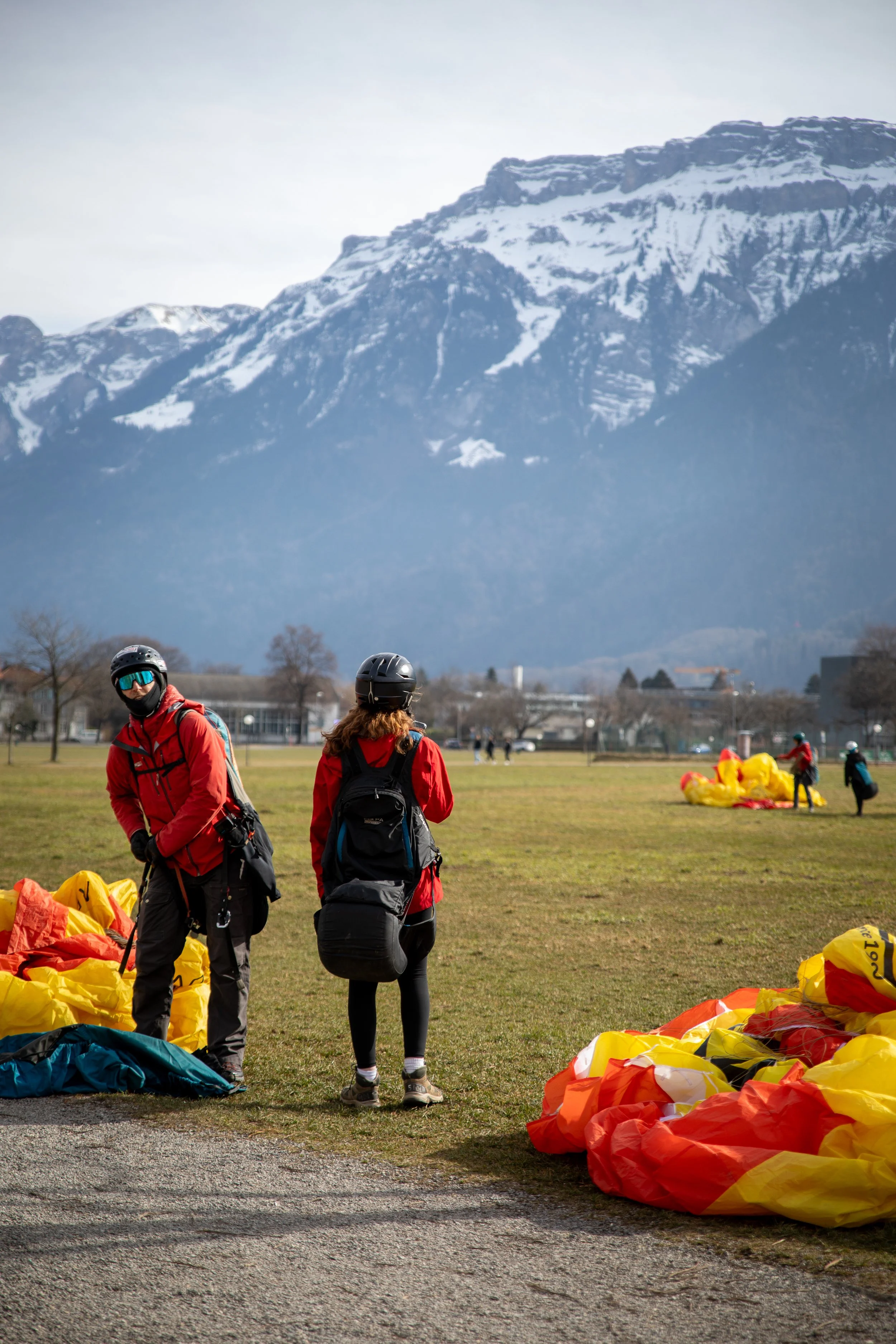 Group of skydivers with gear preparing on a field with snow-capped mountains in the background.